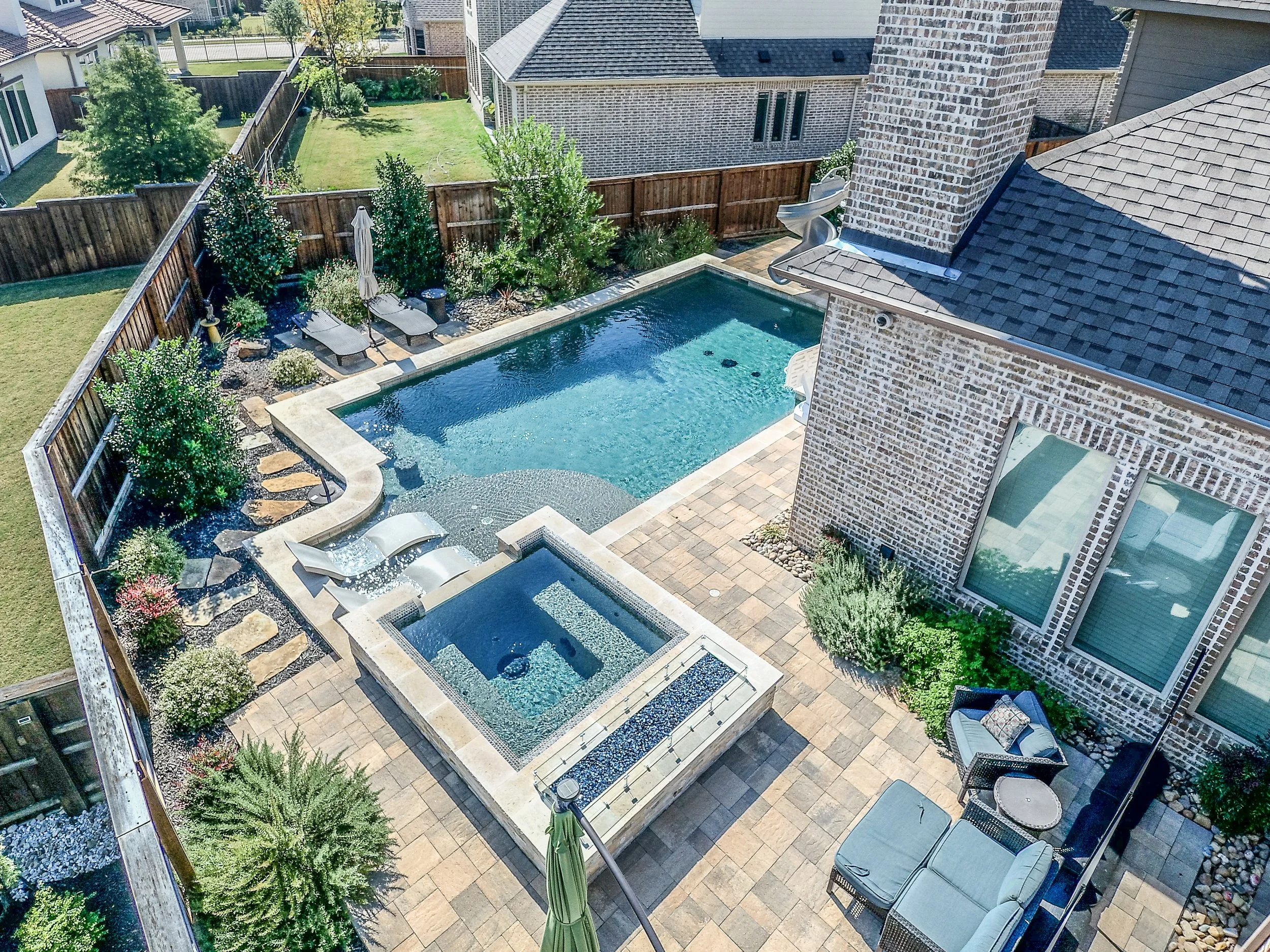 Aerial view of a backyard featuring a swimming pool, a hot tub, and a lounging area with chairs and plants, surrounded by a wooden fence and neighboring houses.