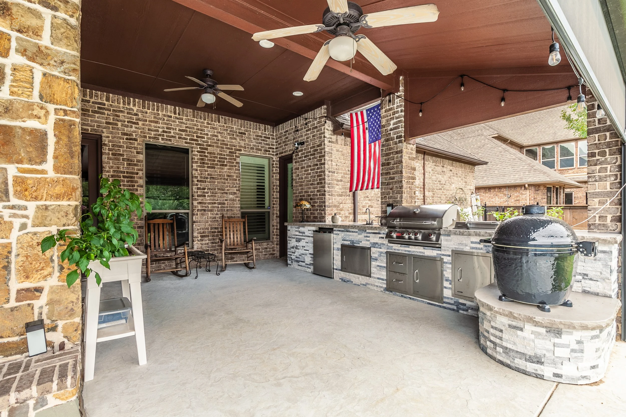 Covered outdoor patio with brick walls, ceiling fans, string lights, a countertop with a grill and a black smoker, two wooden chairs, a potted plant, and an American flag hanging.