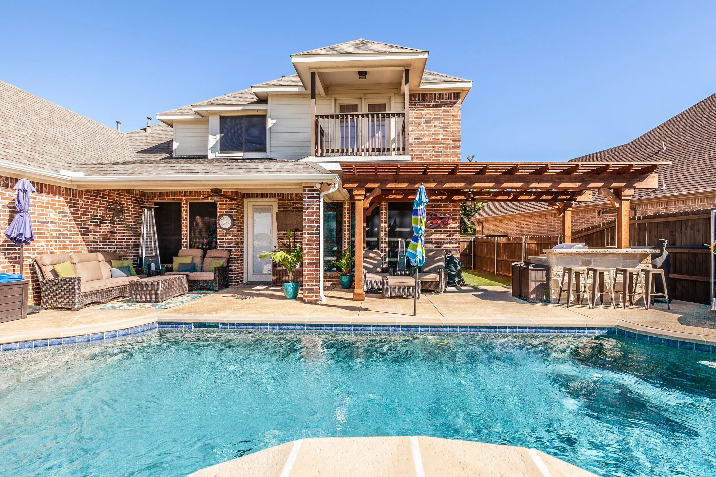 Backyard with swimming pool, patio furniture, and a wooden pergola in front of a brick house with a balcony.