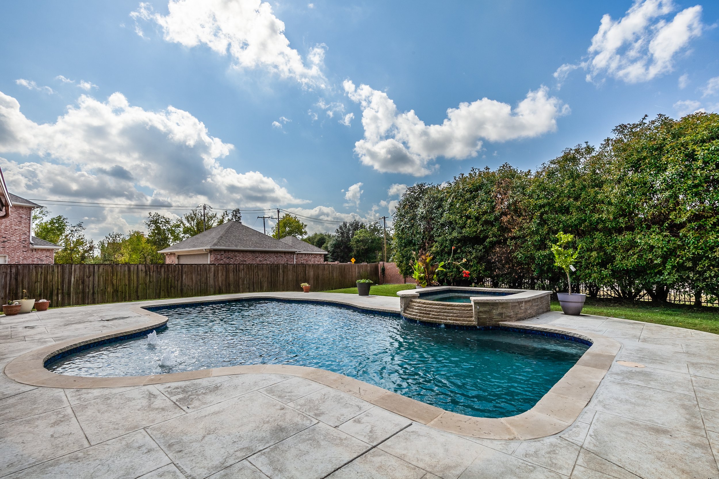 Backyard swimming pool with a hot tub, stone deck, potted plants, a wooden fence, trees, a blue sky with clouds.