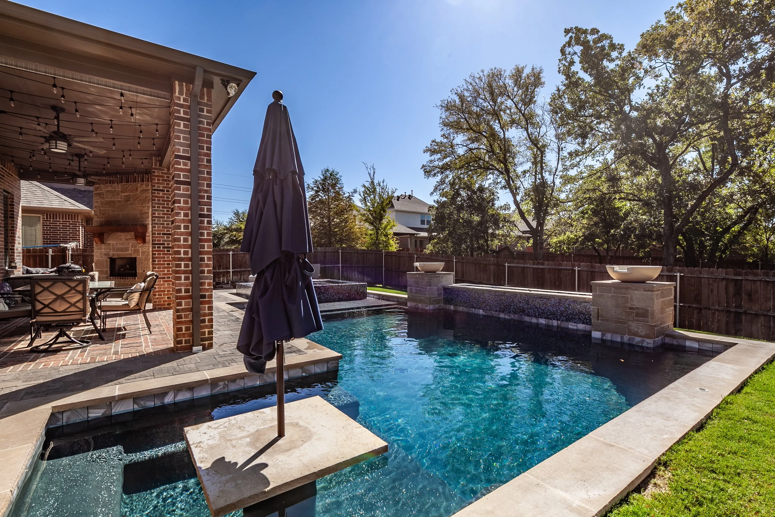 Backyard pool with umbrella, patio furniture, and water features under a clear blue sky, surrounded by trees and a wooden fence.