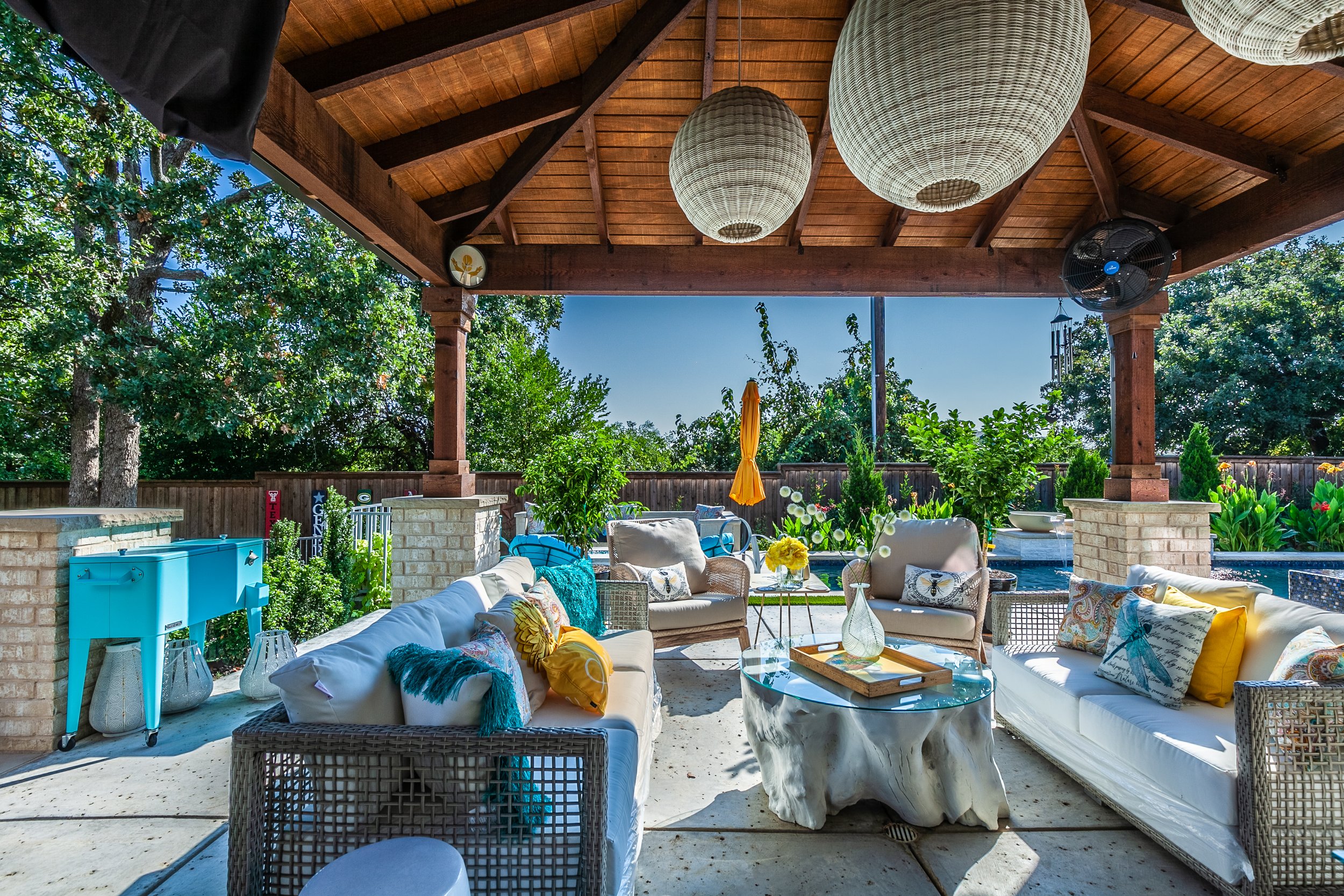 Covered outdoor patio with white sofas, a glass coffee table, and decorative pillows, surrounded by green trees and a wooden fence, with a bright sunny sky.