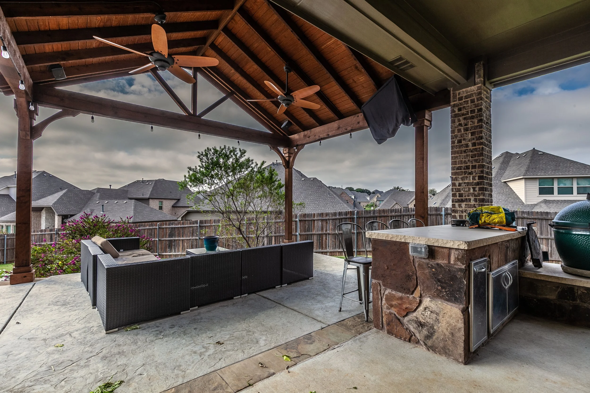 Covered outdoor patio with wooden ceiling and ceiling fans, seating area with black wicker sofa and cushions, a bar with stone and countertop, grill, and a view of neighboring houses and a cloudy sky.