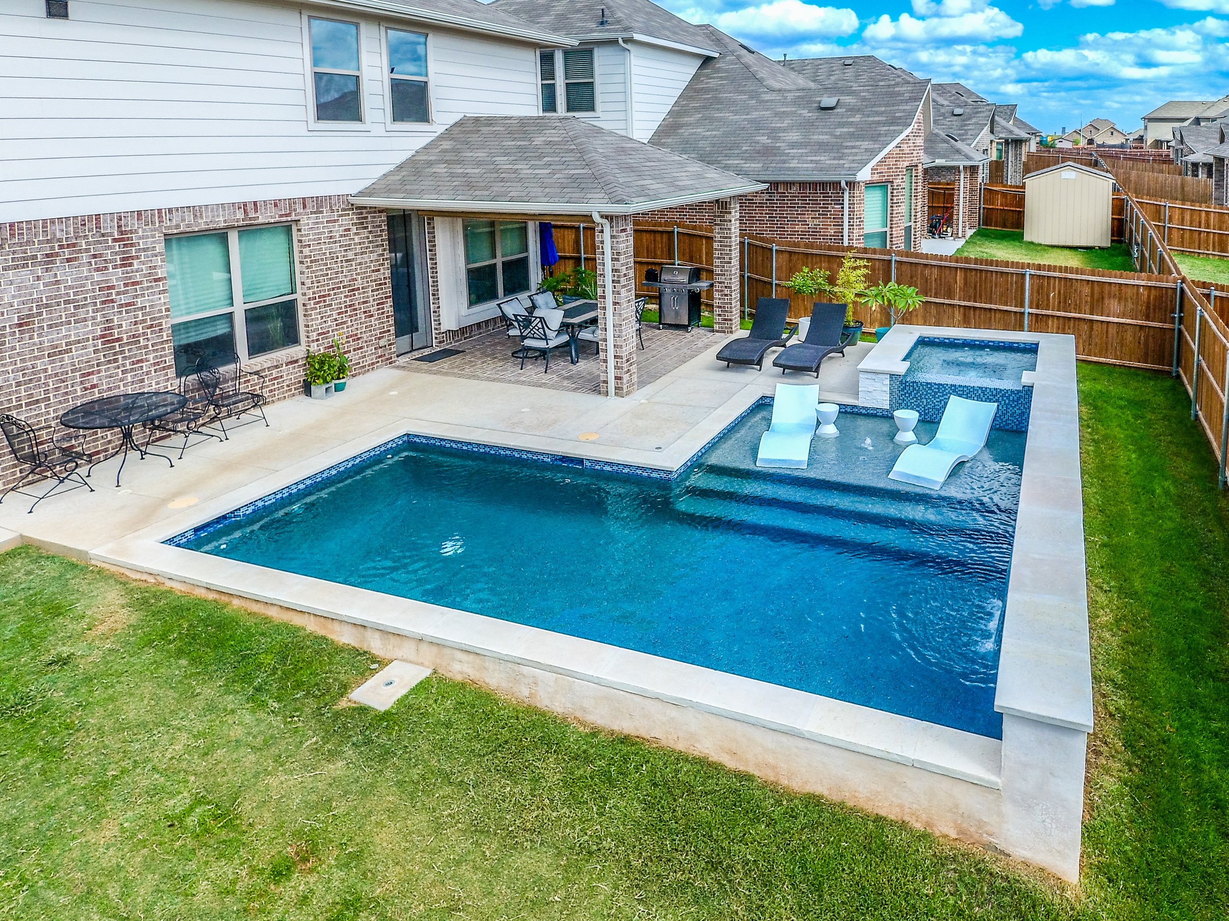 Backyard with a pool and hot tub, outdoor seating, and a covered patio area with a grill, surrounded by a wooden fence.