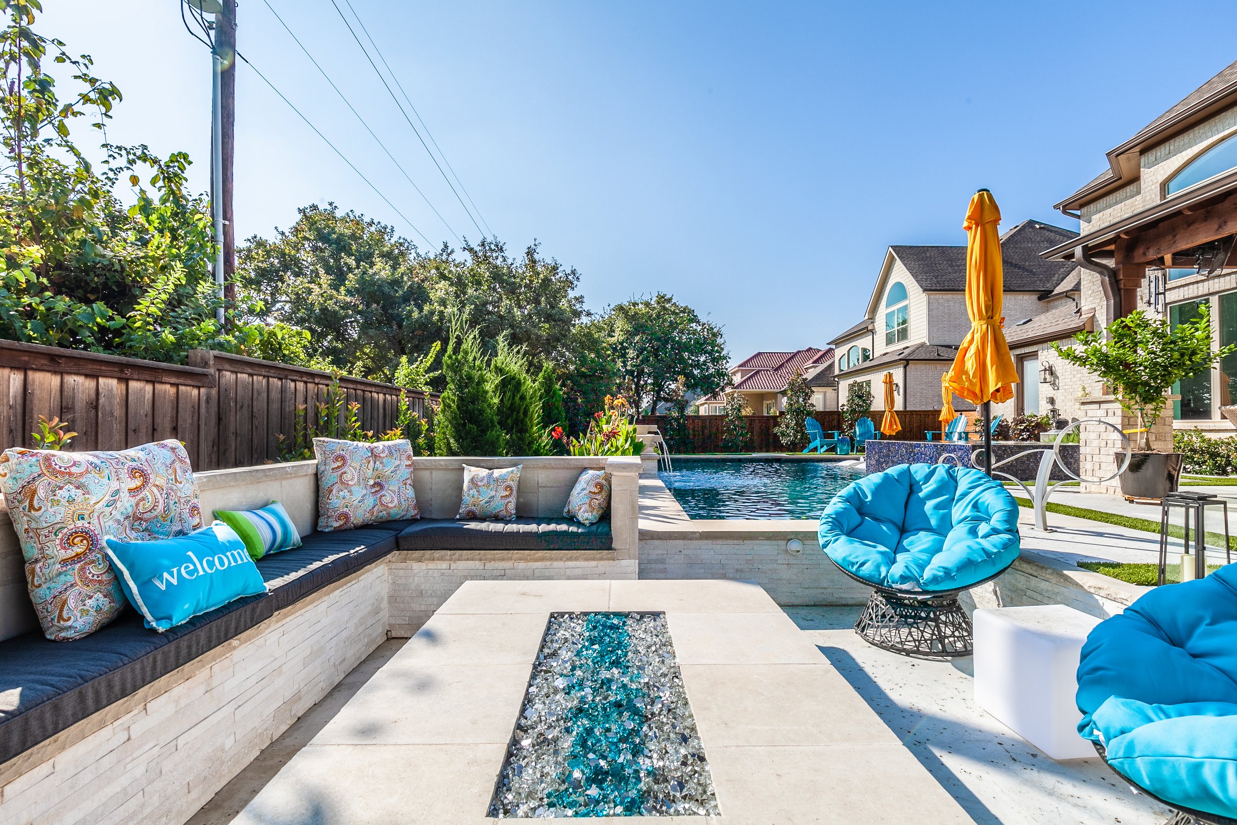 Backyard patio with swimming pool, colorful cushions, blue chairs with cushions, yellow umbrellas, and greenery with houses in the background