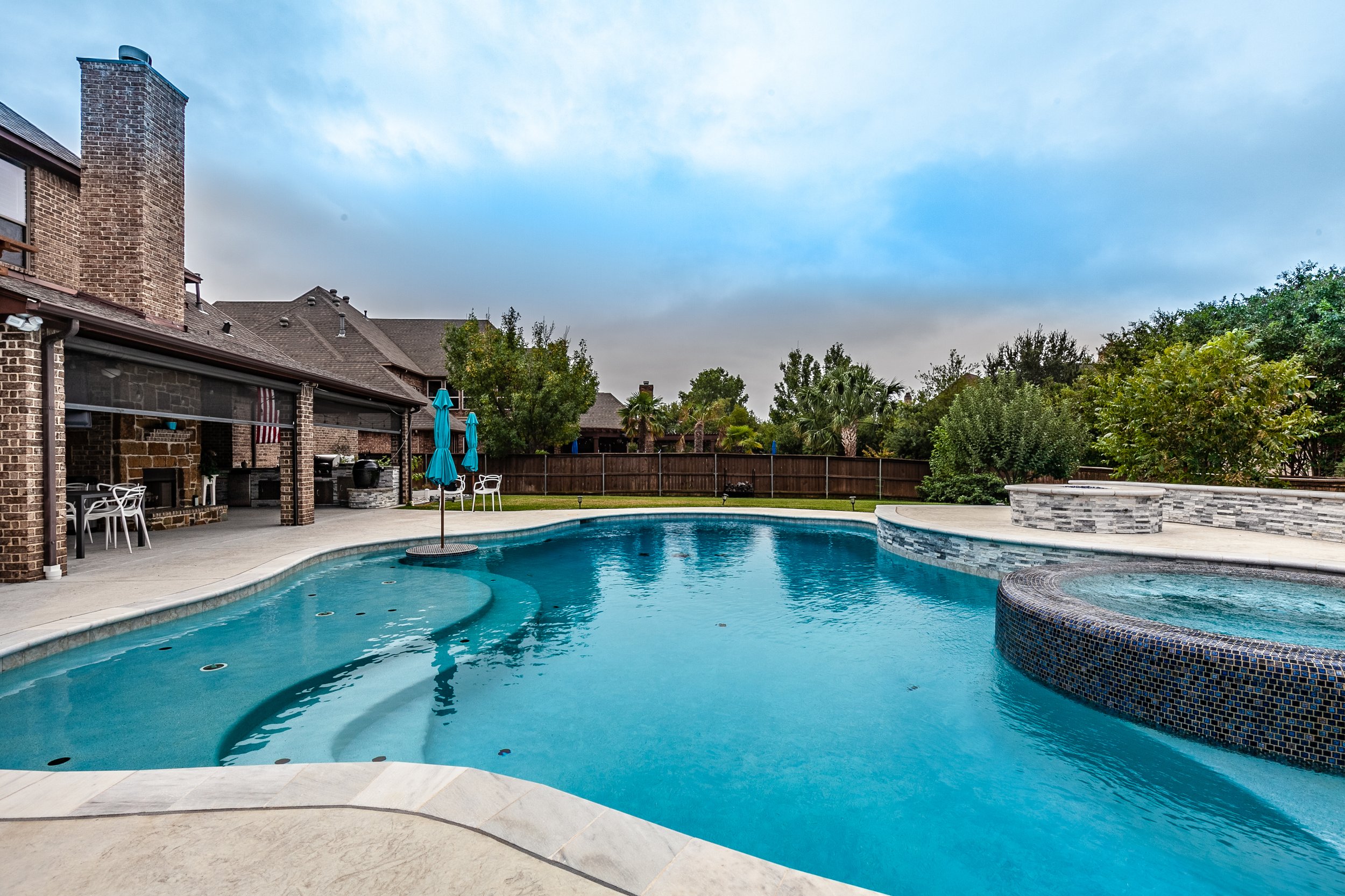 Backyard with a swimming pool, blue umbrellas, and lounge chairs near a brick house with a chimney, surrounded by trees and a wooden fence.
