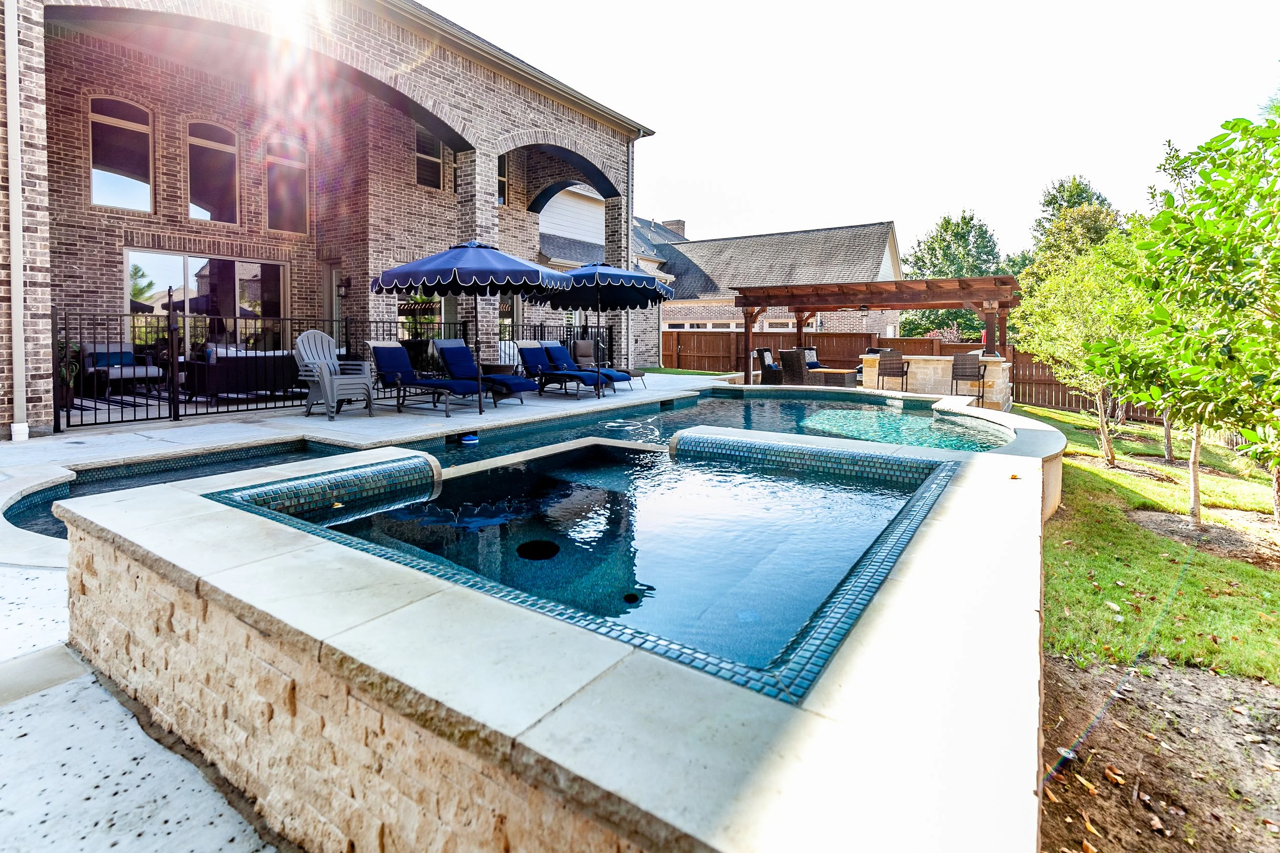 Backyard with swimming pool, lounge chairs under umbrellas, and a brick house in the background.