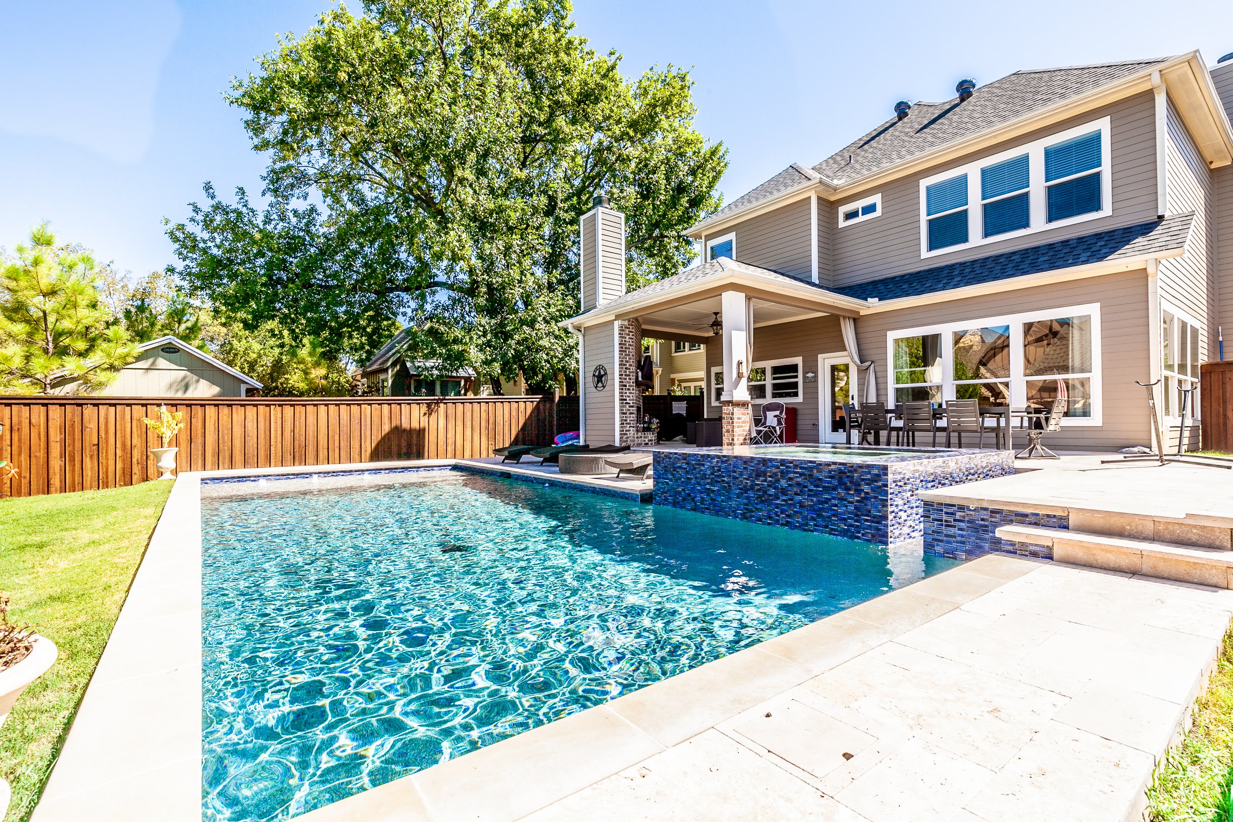 A backyard with a swimming pool, wooden fence, trees, and a two-story house with large windows and a covered patio with outdoor furniture.