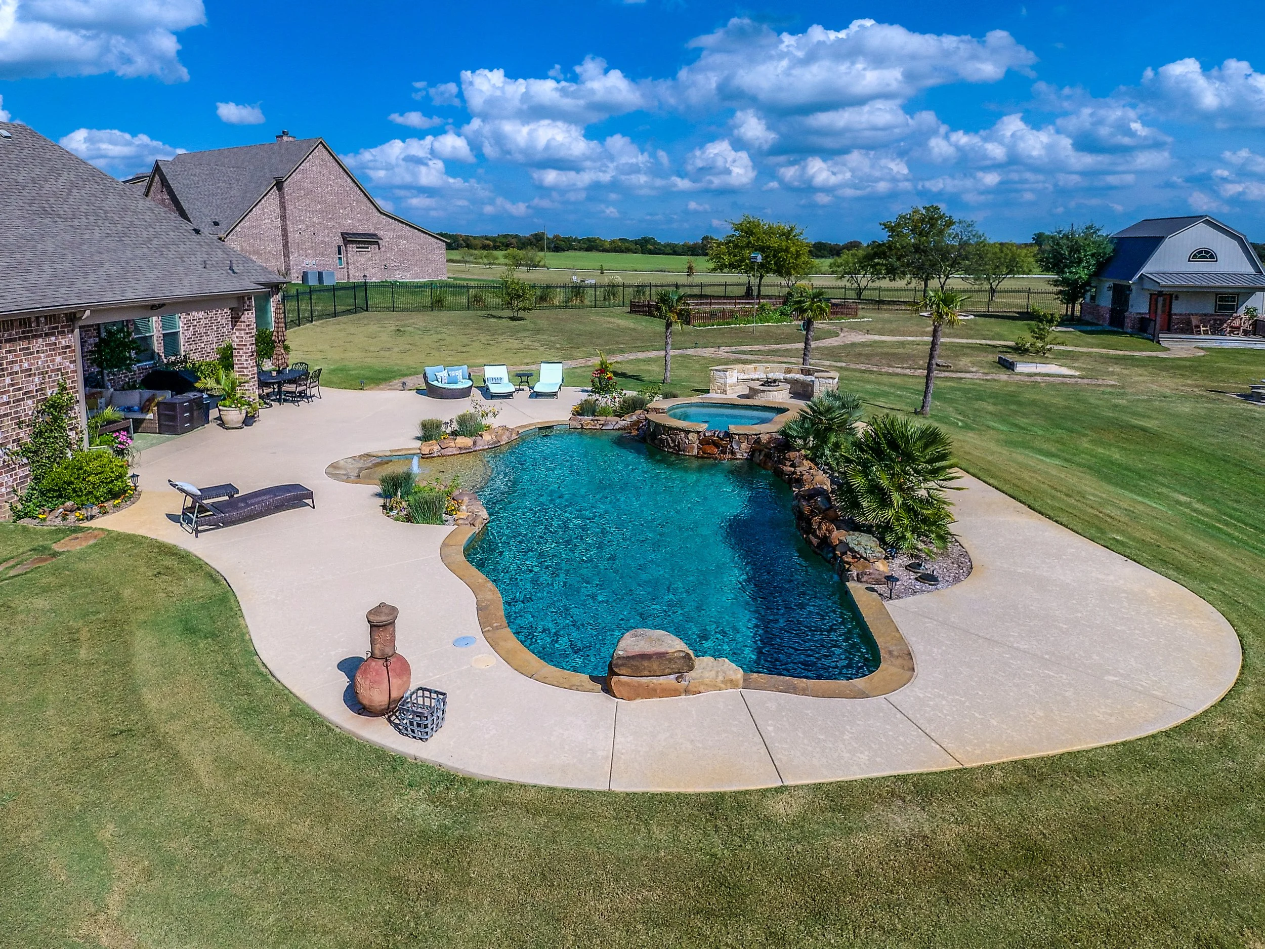 A backyard with a swimming pool surrounded by a concrete patio, featuring lounge chairs, potted plants, and outdoor seating. The yard has a well-maintained lawn, palm trees, and a hot tub, with a brick house and additional structures in the backgroun