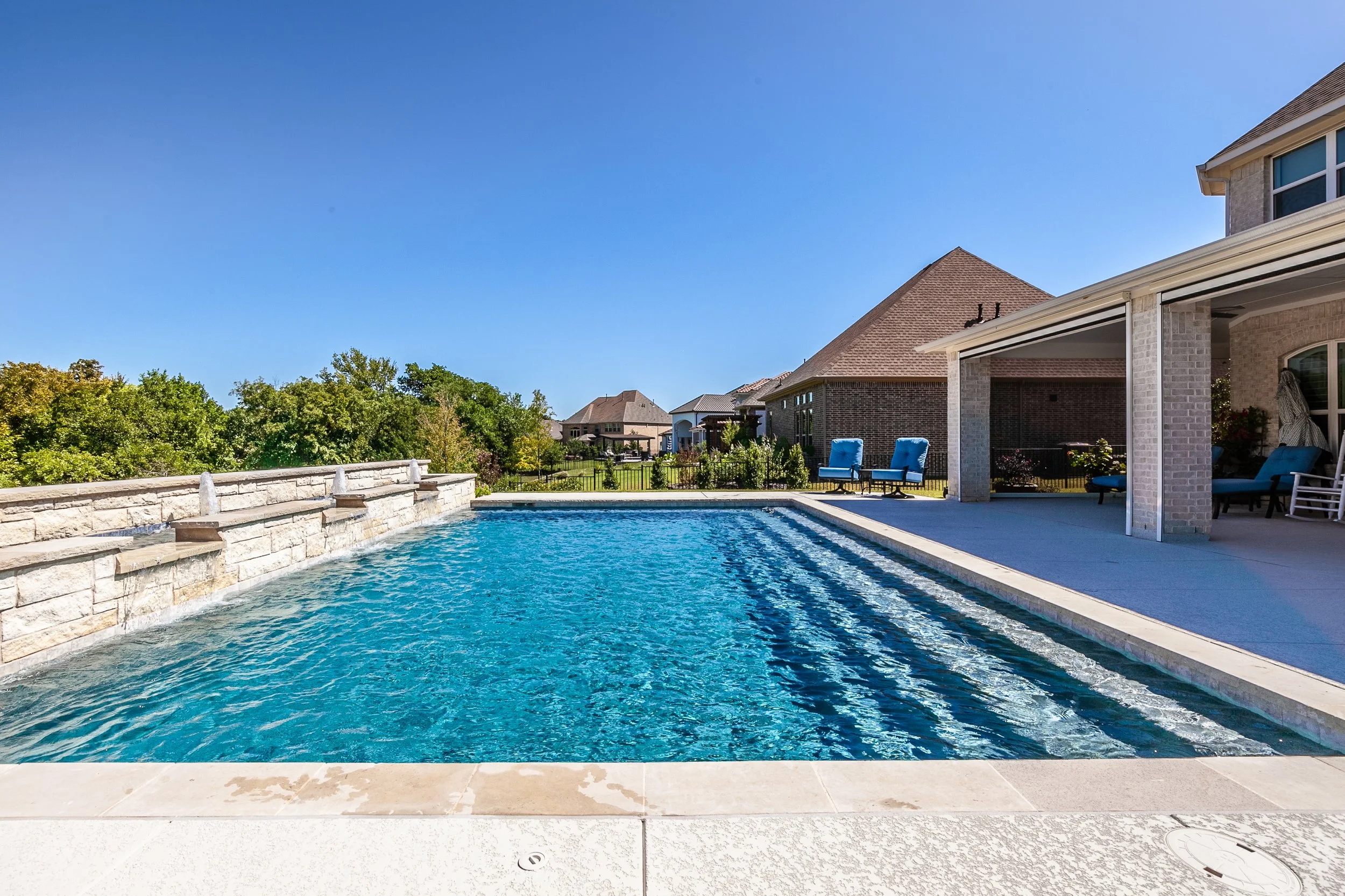 A backyard swimming pool with water flowing from a stone fountain, surrounded by a concrete deck with outdoor furniture, under a clear blue sky.
