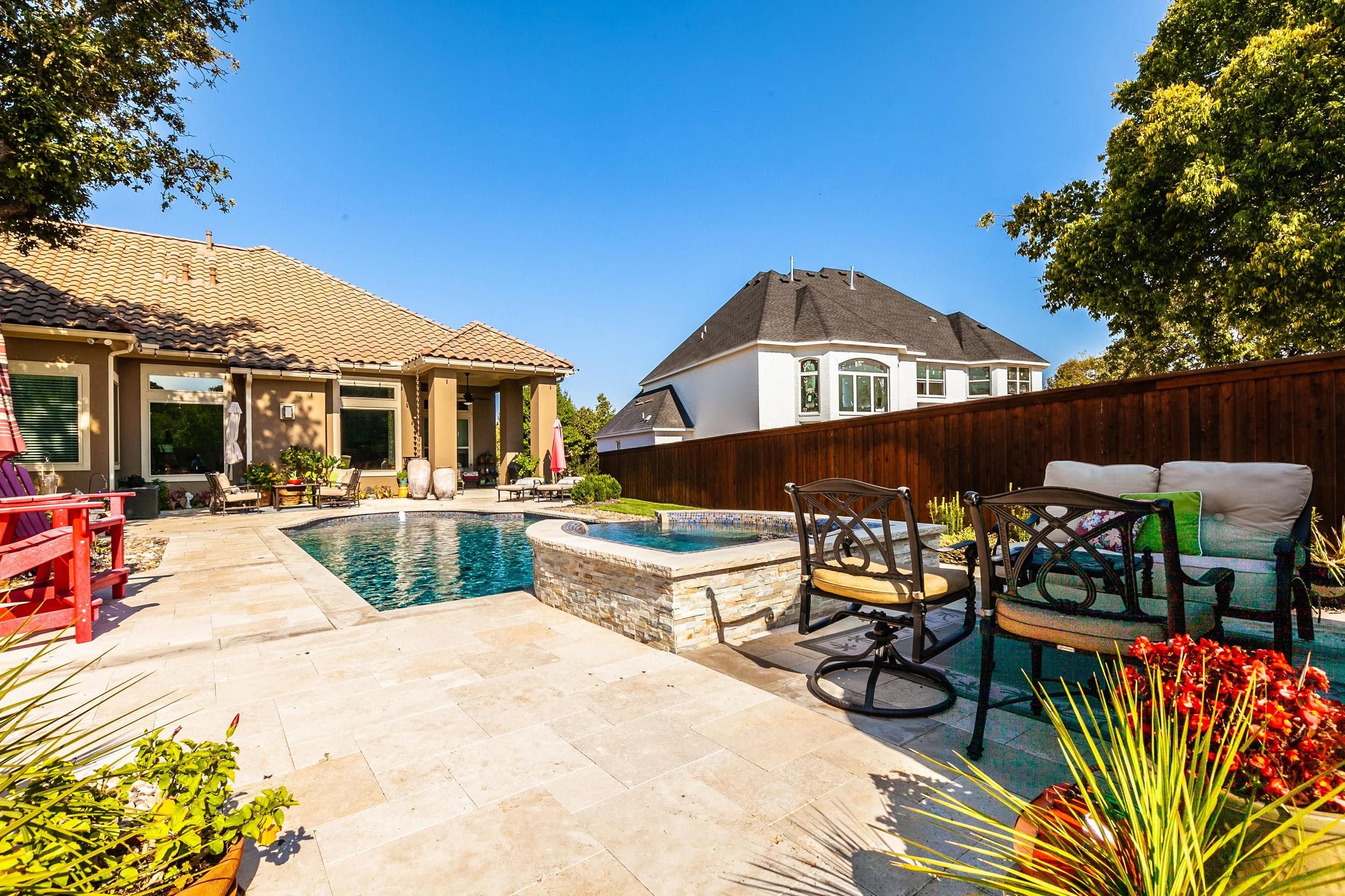 A backyard with a swimming pool, hot tub, patio furniture, and a wooden fence under a clear blue sky.