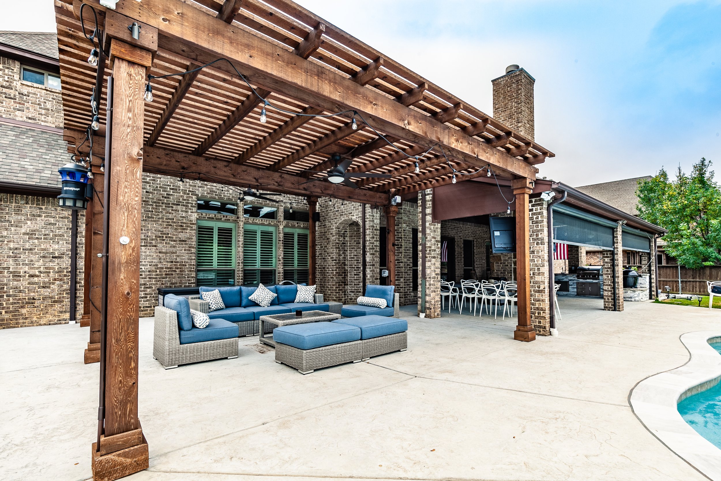 Backyard patio area featuring a wooden pergola with hanging string lights, outdoor seating with blue cushions, a grill area, and a swimming pool.