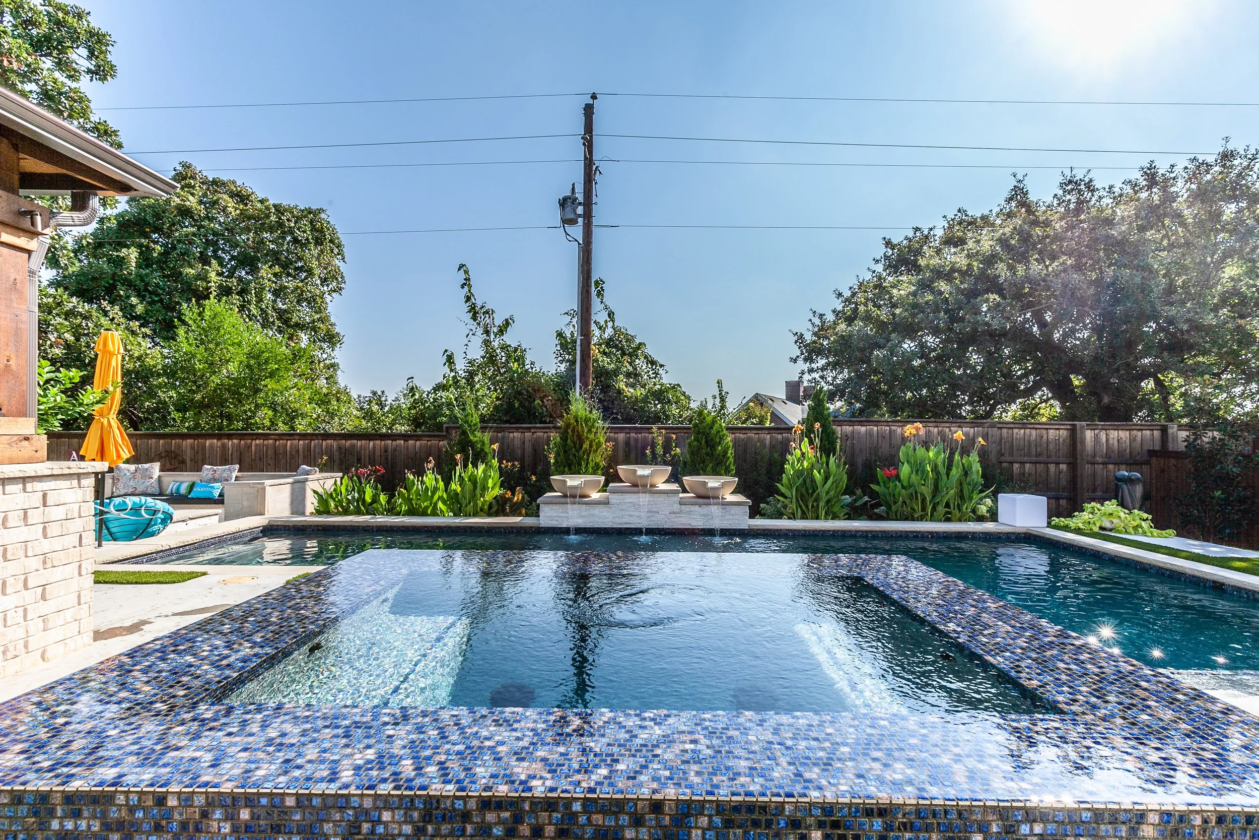 Residential backyard with a swimming pool, surrounded by greenery, a wooden fence, and outdoor seating area with yellow umbrella and cushions, under a clear sunny sky.