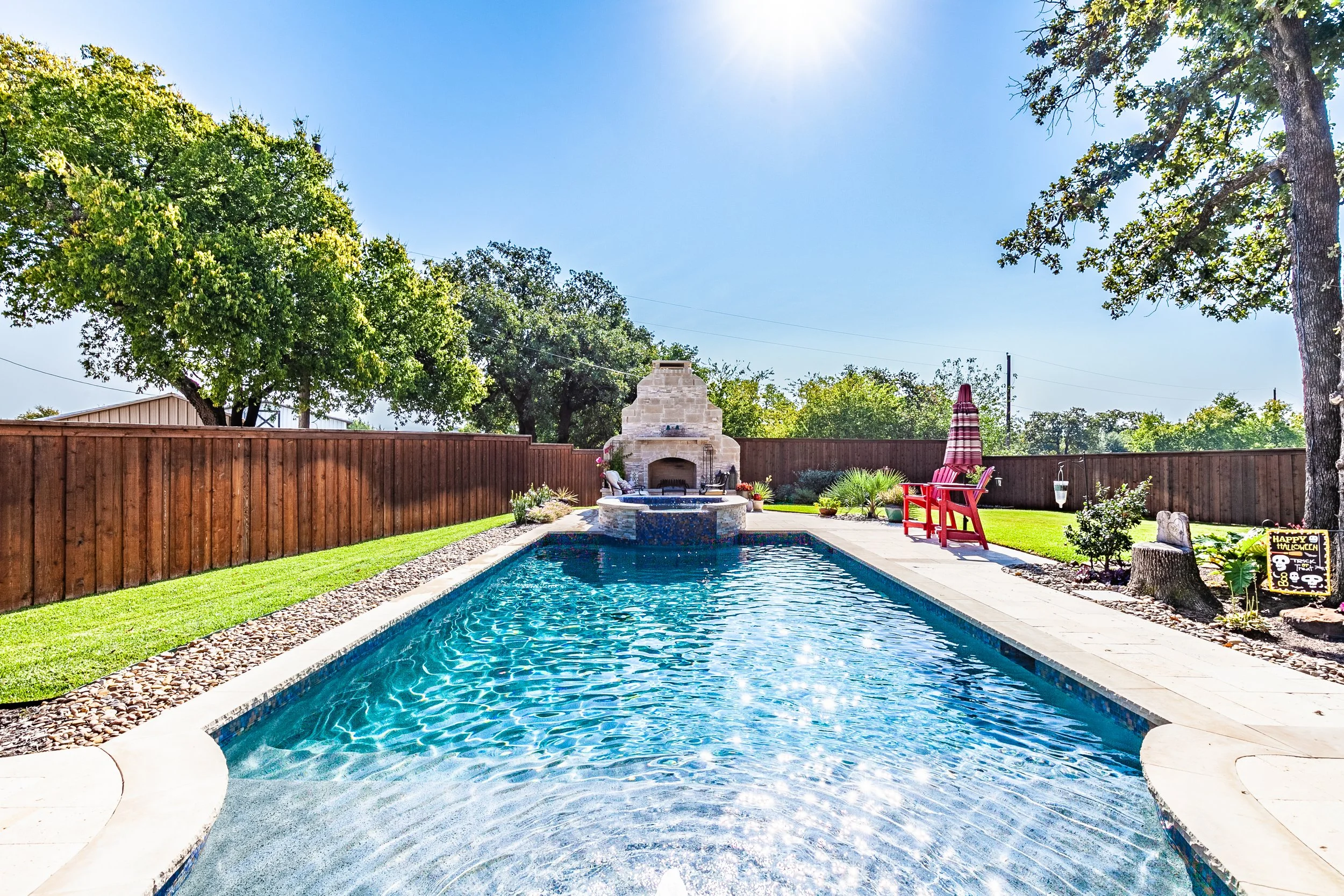 A backyard swimming pool with a stone fireplace at the far end, surrounded by green grass, trees, and a fenced boundary. A red chair with a striped umbrella is on the right side, along with decorative plants, a tree stump, and Halloween-themed decora