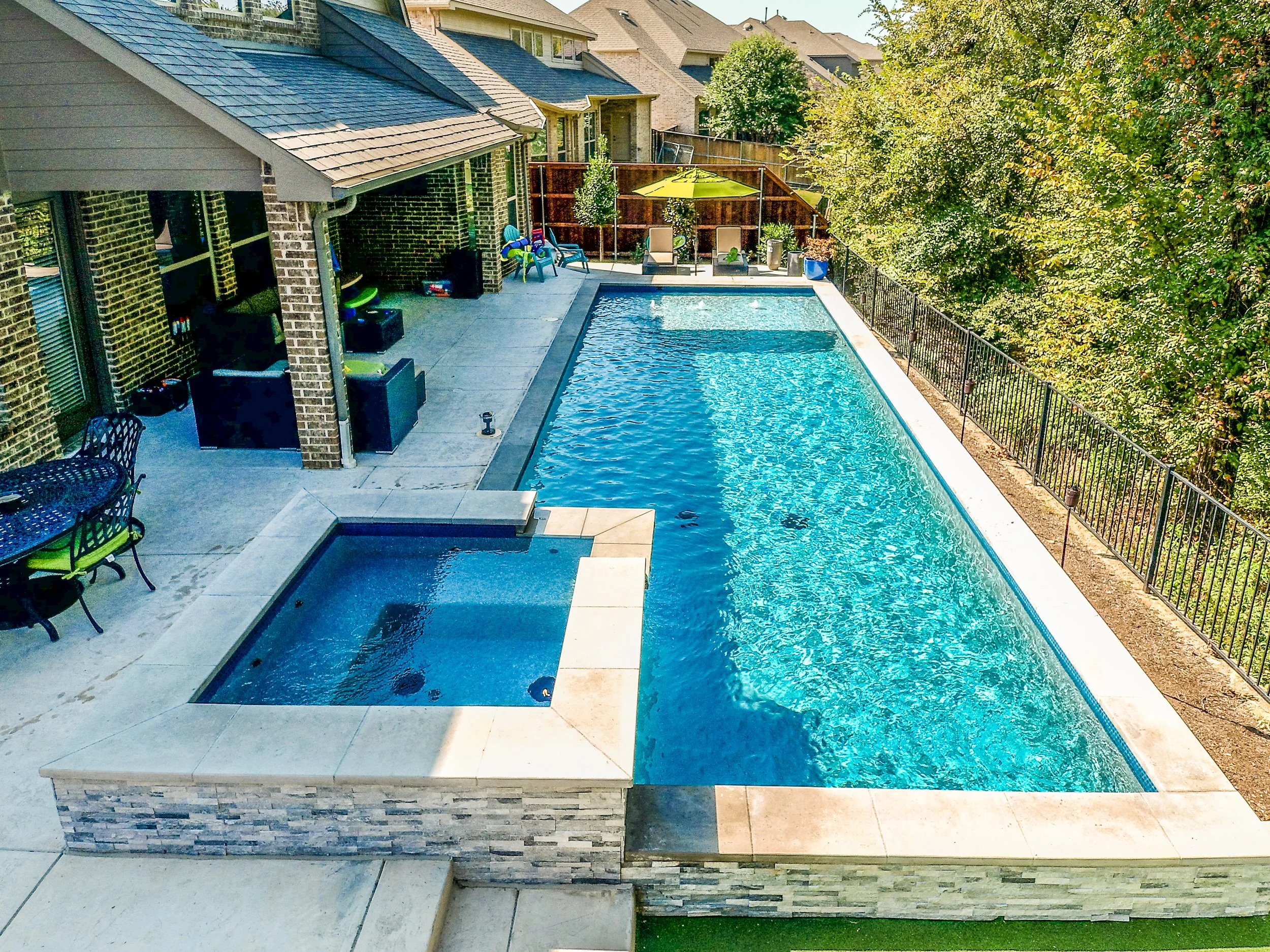 A backyard swimming pool with a hot tub, patio furniture, and trees surrounding the area.
