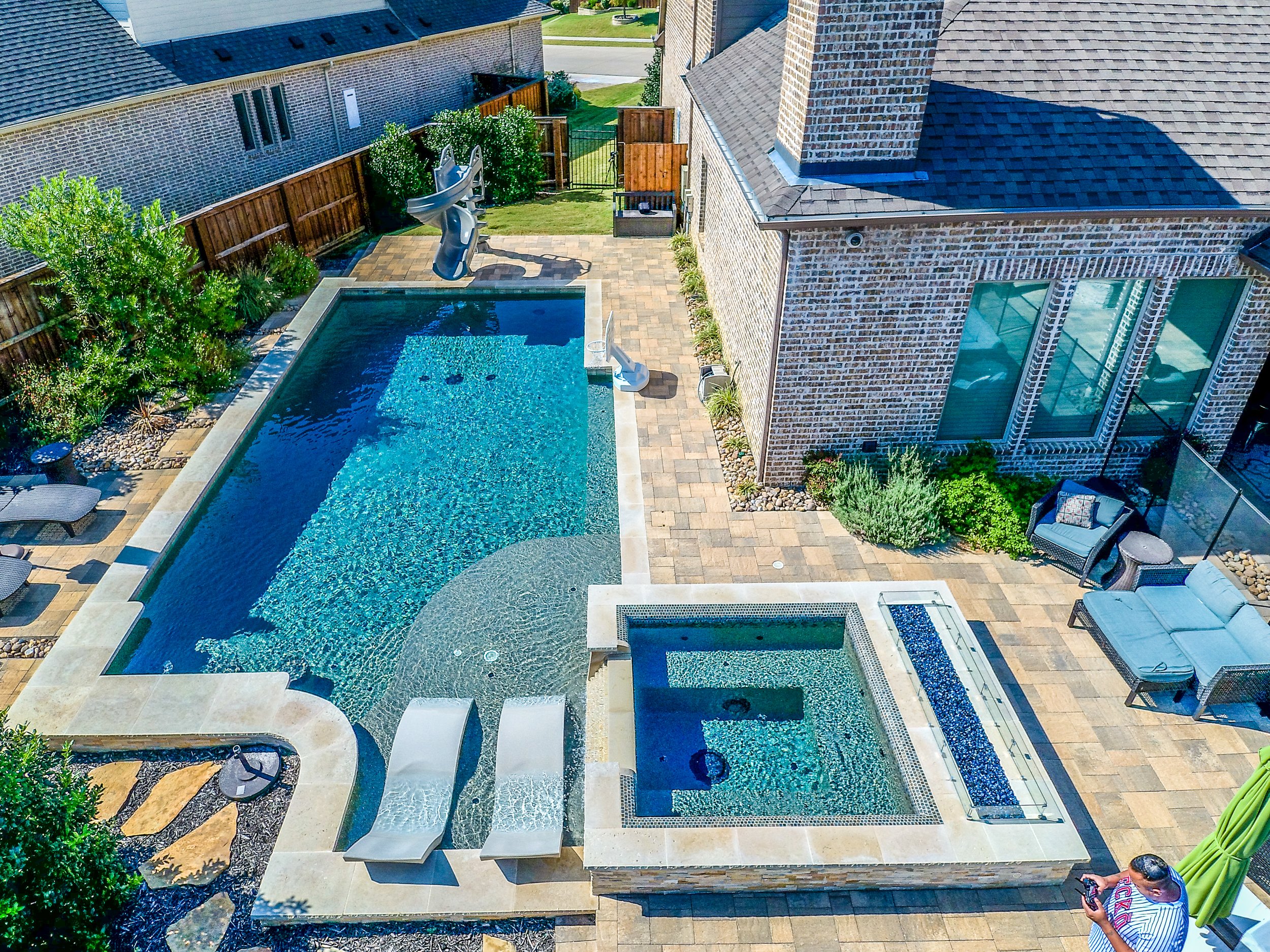 Overhead view of a backyard with a swimming pool and hot tub, surrounded by patio furniture, a slide, shrubs, and a brick house with large windows.