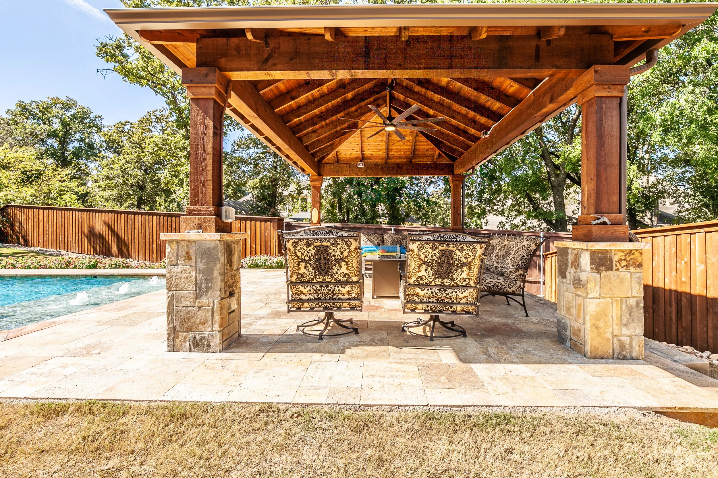 Am outdoor covered patio area with stone columns, a wooden roof with a ceiling fan, and a patio table with six ornate chairs, next to a swimming pool and surrounded by trees and a wooden fence.