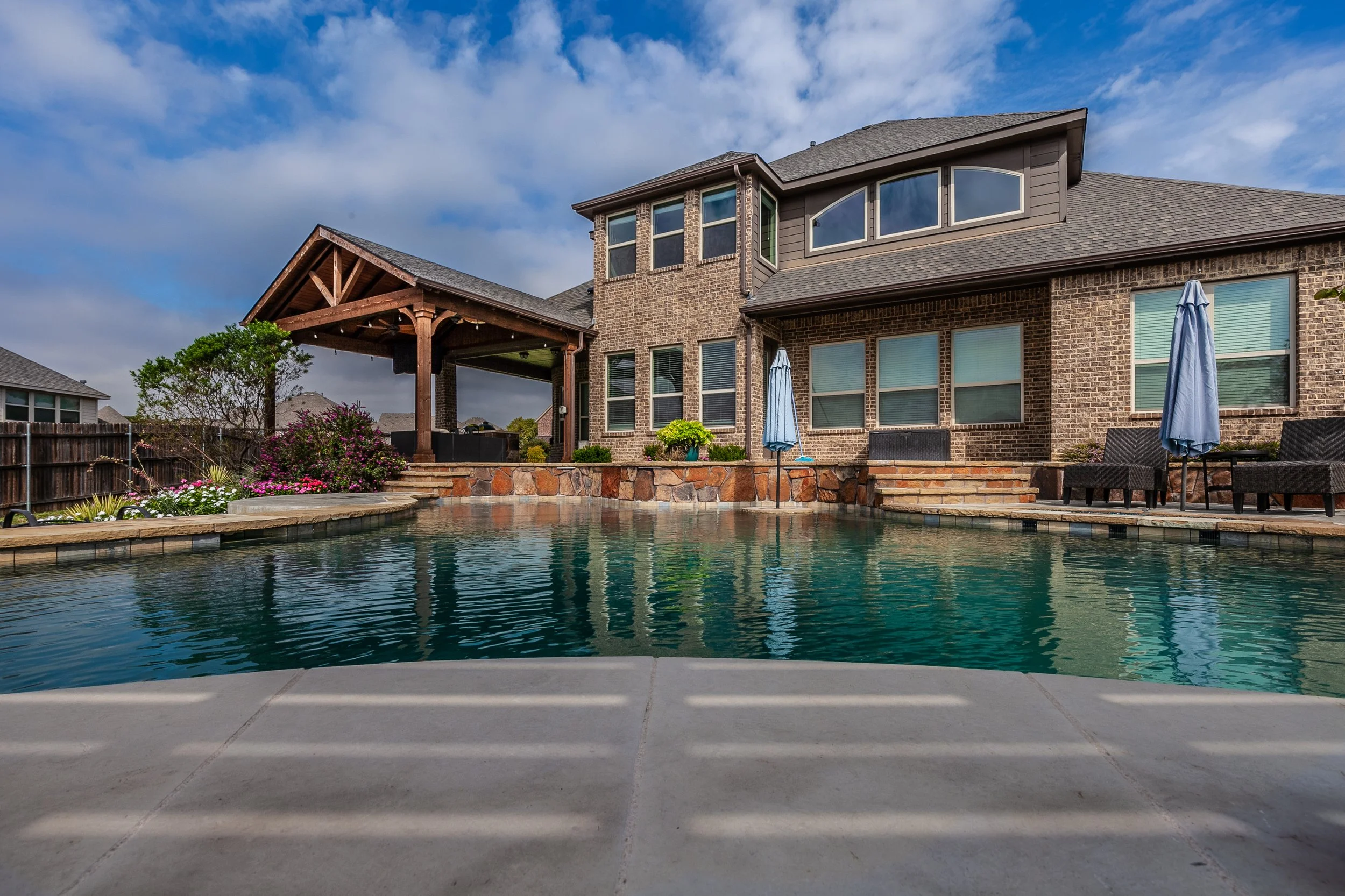 Backyard with a swimming pool, patio chairs with umbrellas, a brick house with multiple windows, and a wooden pergola under a partly cloudy sky.