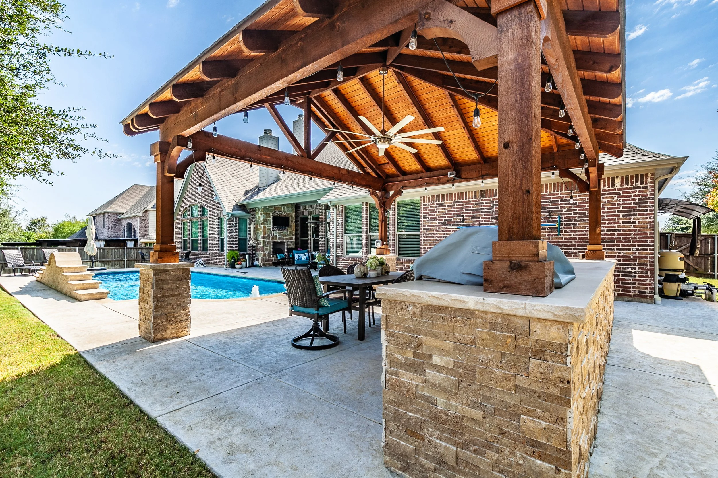 Luxury backyard with swimming pool, stone and brick house, covered patio with a ceiling fan, outdoor furniture, and string lights.