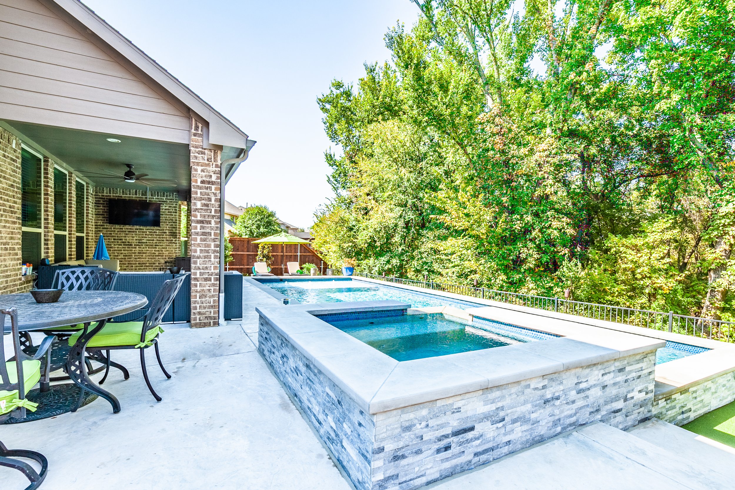 Backyard patio with swimming pool and hot tub, outdoor furniture, trees, and blue sky.