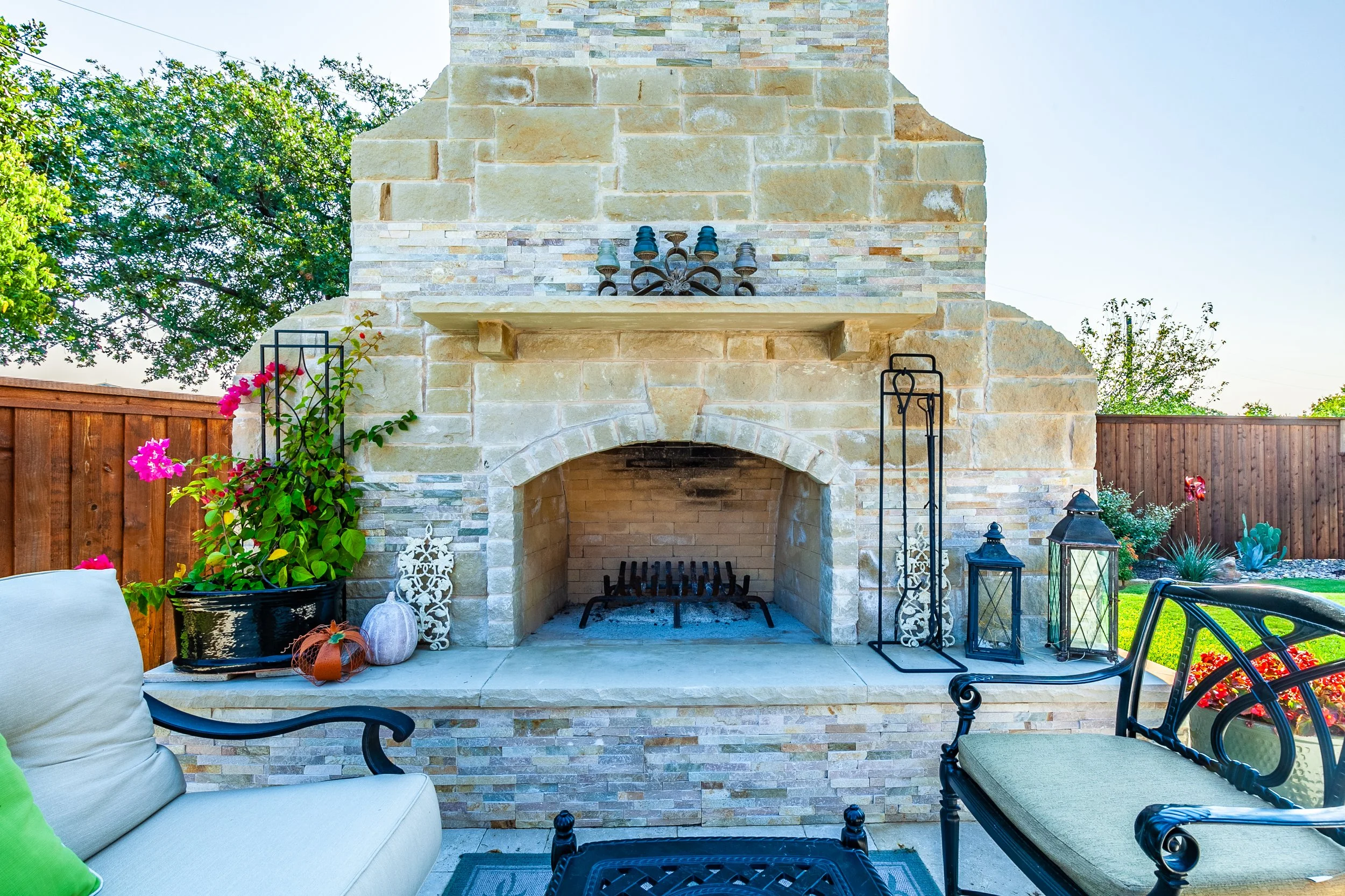 Outdoor patio area with a stone fireplace, black metal and glass lanterns, potted plants with pink and orange flowers, seating with cushions, and a wooden fence in the background.