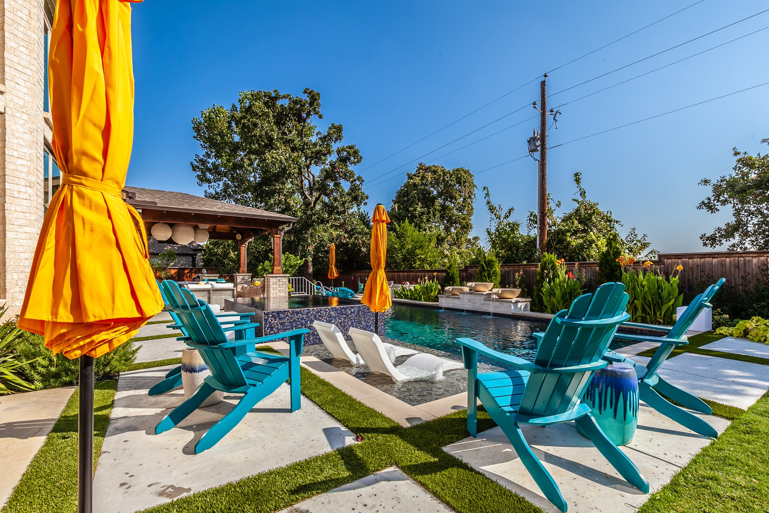 A backyard with a swimming pool, four blue Adirondack chairs, two white lounge chairs, yellow umbrellas, a pergola with decorative lanterns, and lush green trees.