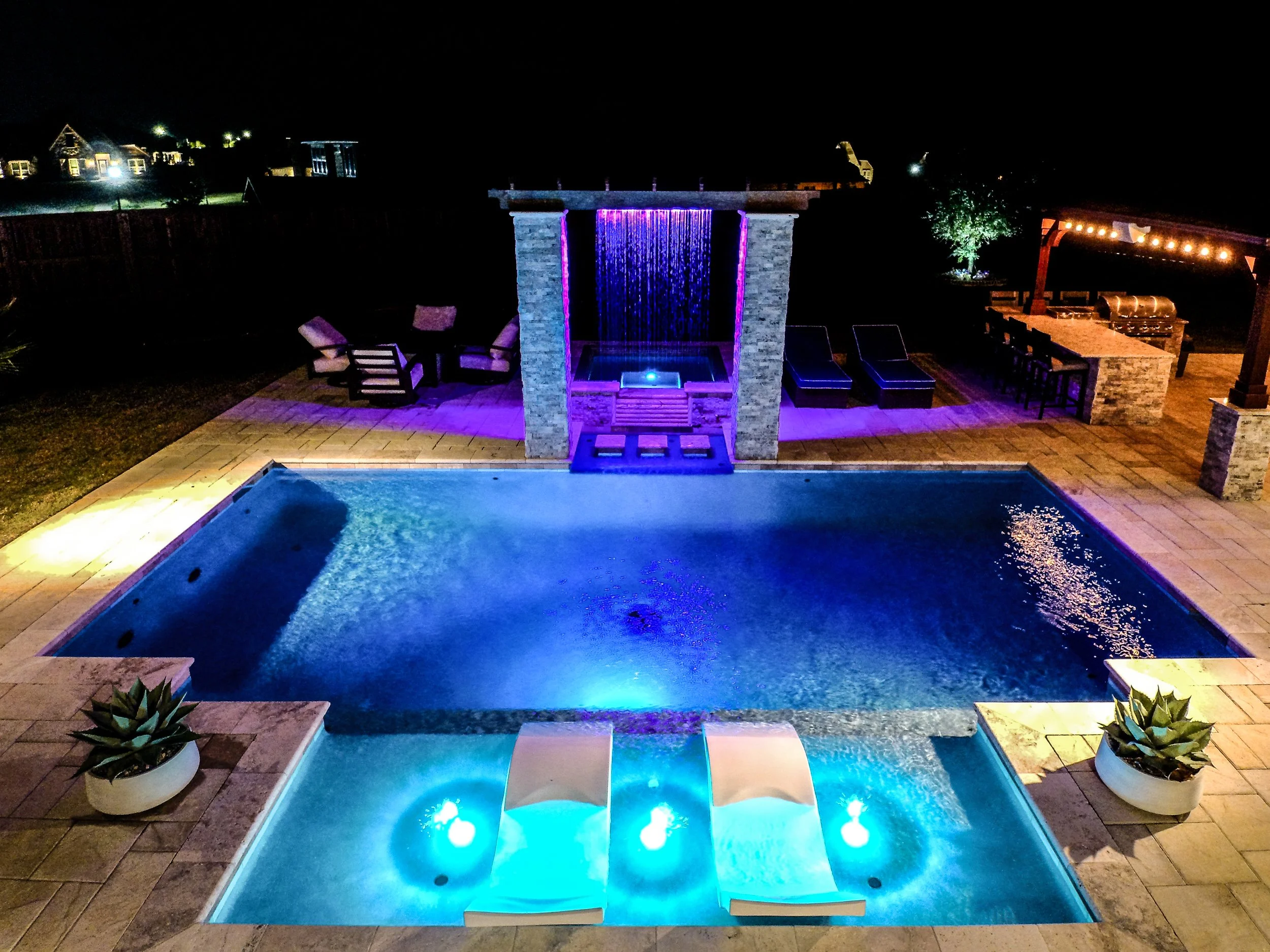 Nighttime view of an illuminated swimming pool with blue lights, surrounded by a stone patio, seating area, waterfall feature, bar area, and potted succulents, with outdoor lighting and a dark sky.