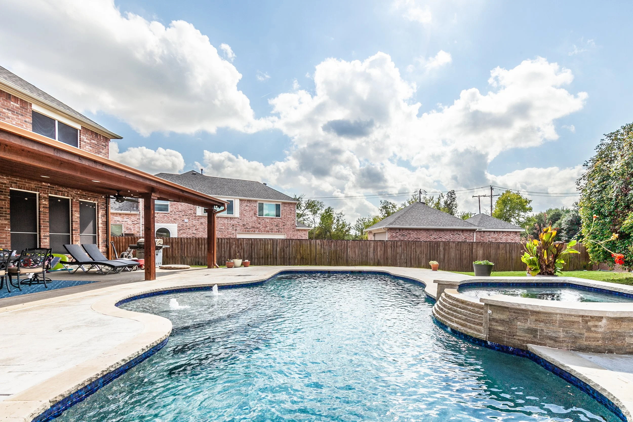 Backyard with a swimming pool, hot tub, patio furniture, and a wooden fence under a partly cloudy sky.