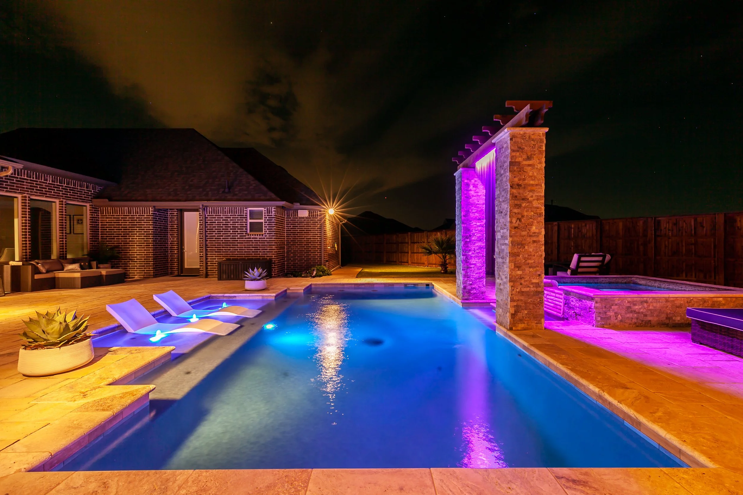 A backyard swimming pool illuminated with colorful underwater and side lighting at night, with lounge chairs and plants around it, and a brick wall and house in the background.