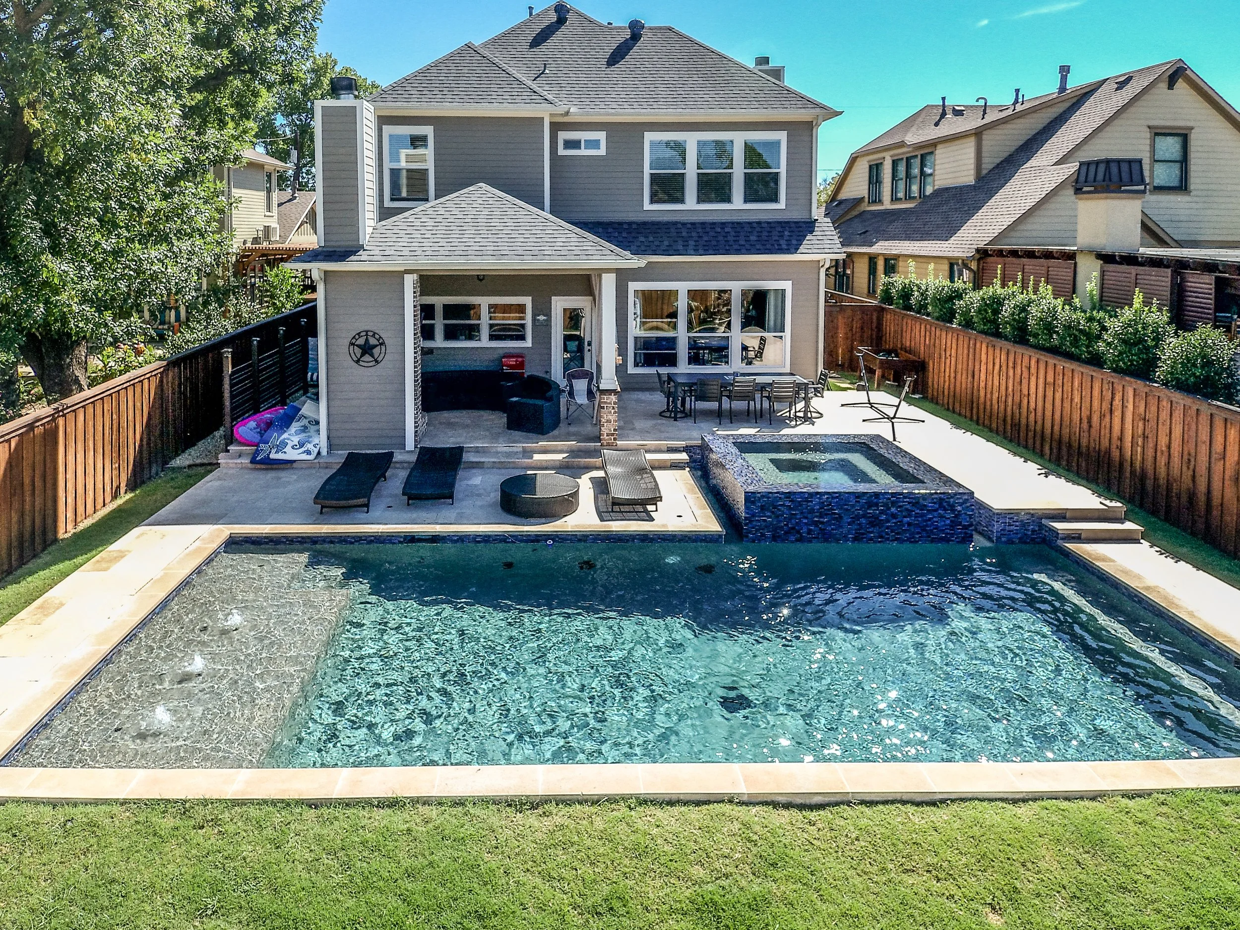 Backyard with a swimming pool, hot tub, and patio seating area, surrounded by a wooden fence and neighboring houses.