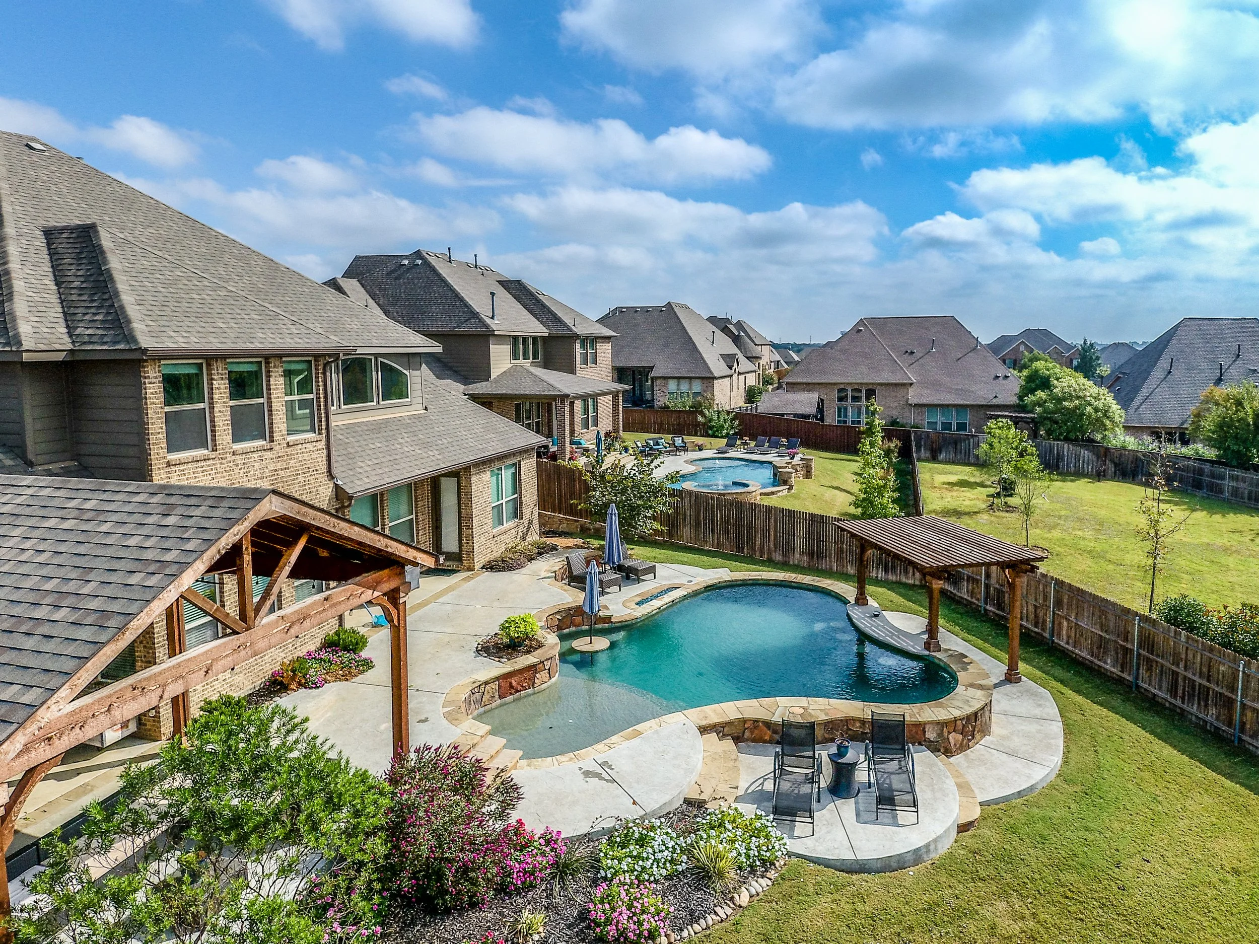 Backyard with a swimming pool, hot tub, covered seating area, lawn, trees, and neighboring houses under a cloudy sky.