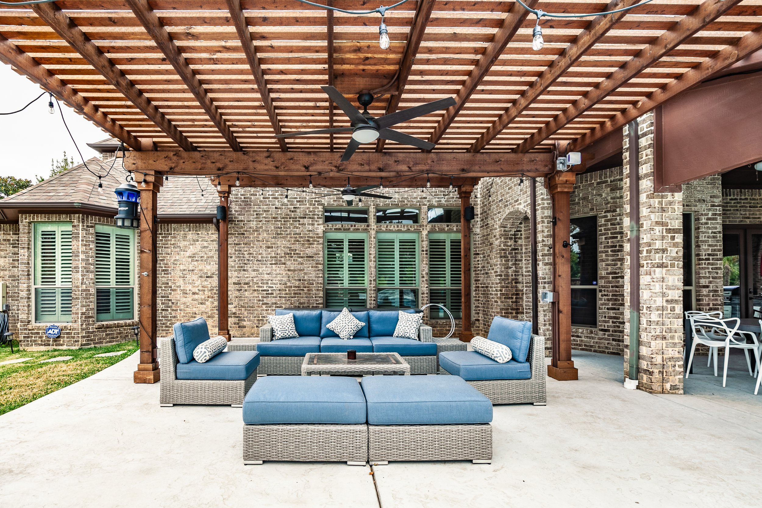 Outdoor patio with wicker furniture and blue cushions, brick house background, wooden pergola with ceiling fans and string lights, and sliding glass door.