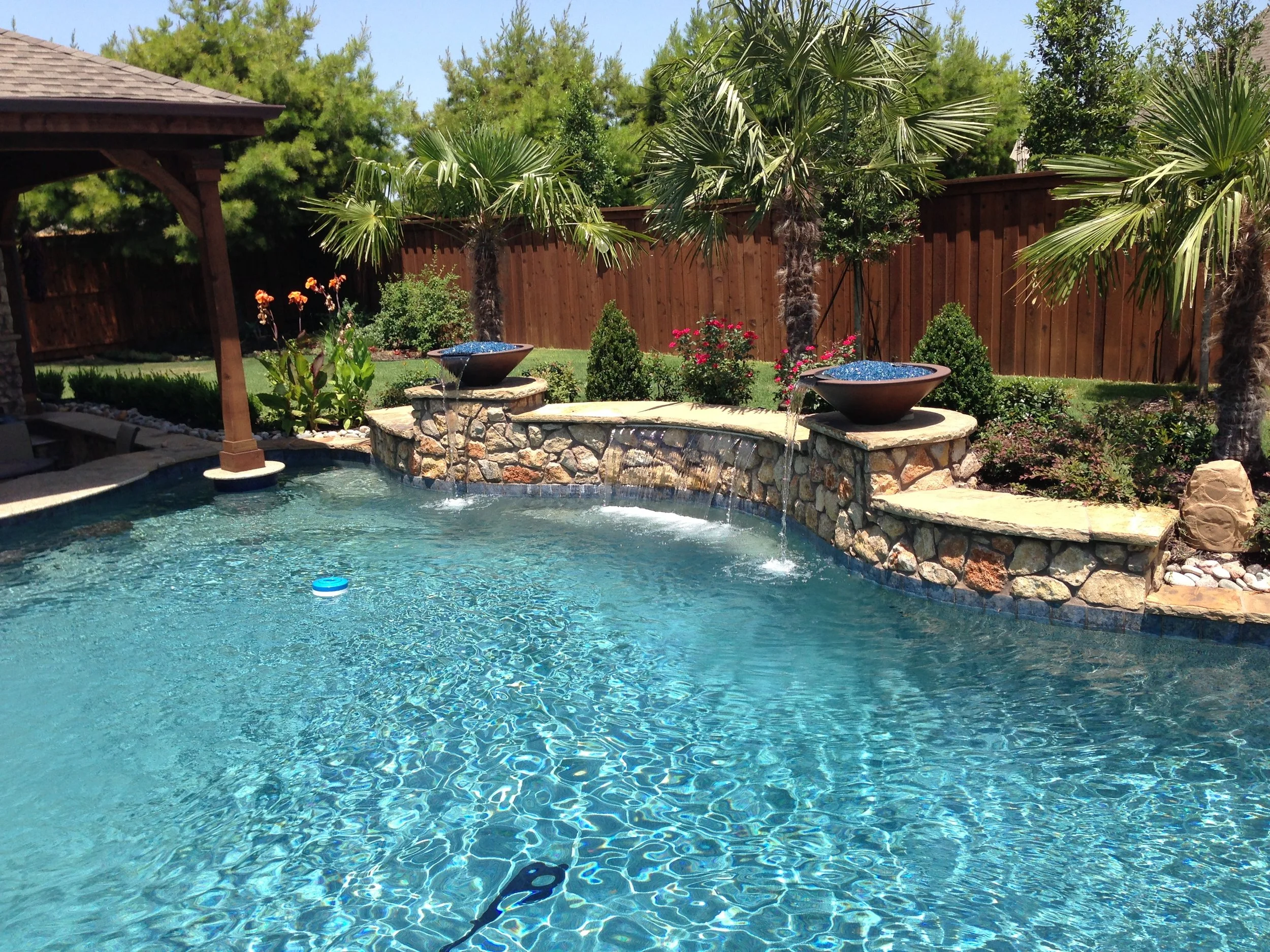 Backyard swimming pool with a stone fountain feature, surrounded by palm trees and various plants, with a wooden privacy fence in the background.