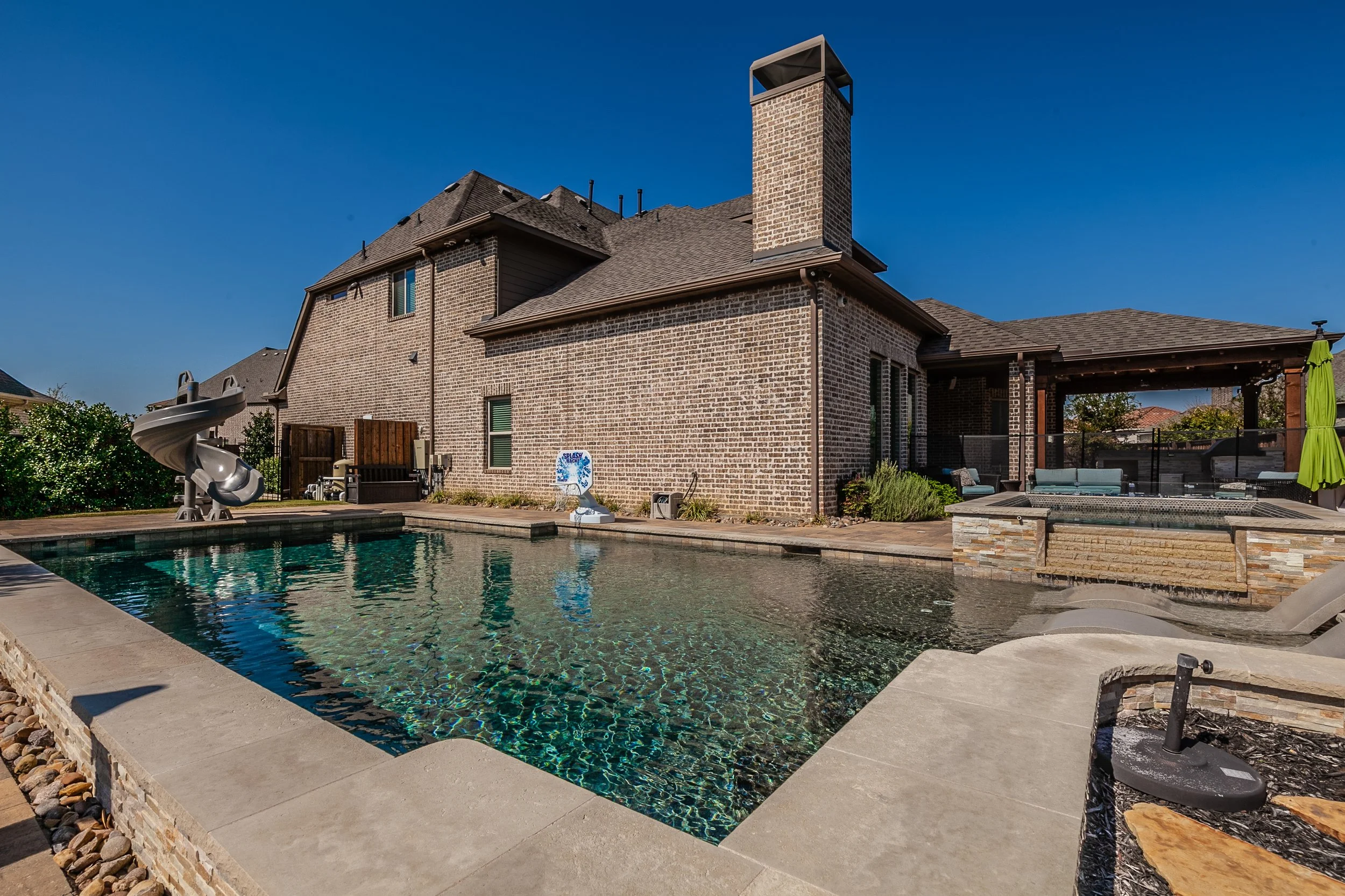 Backyard with a swimming pool, water slide, and an outdoor seating area under a covered patio, with a brick house and a blue sky in the background.