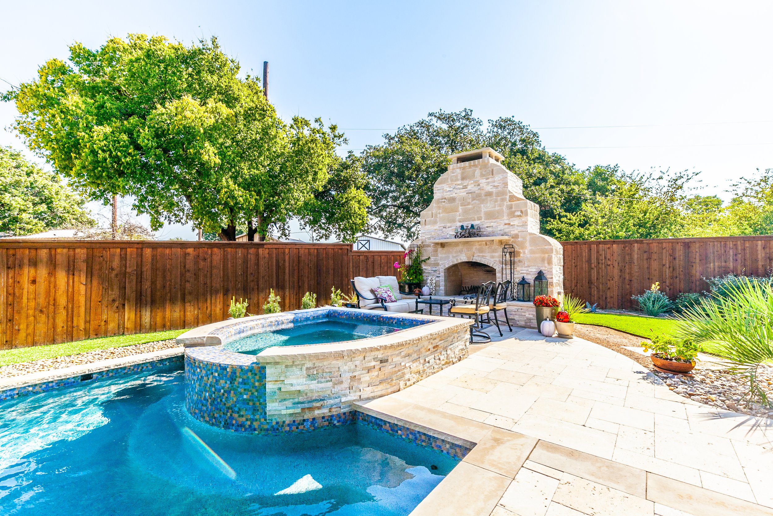 Backyard with swimming pool, jacuzzi, stone fireplace, patio furniture, and garden during daytime.