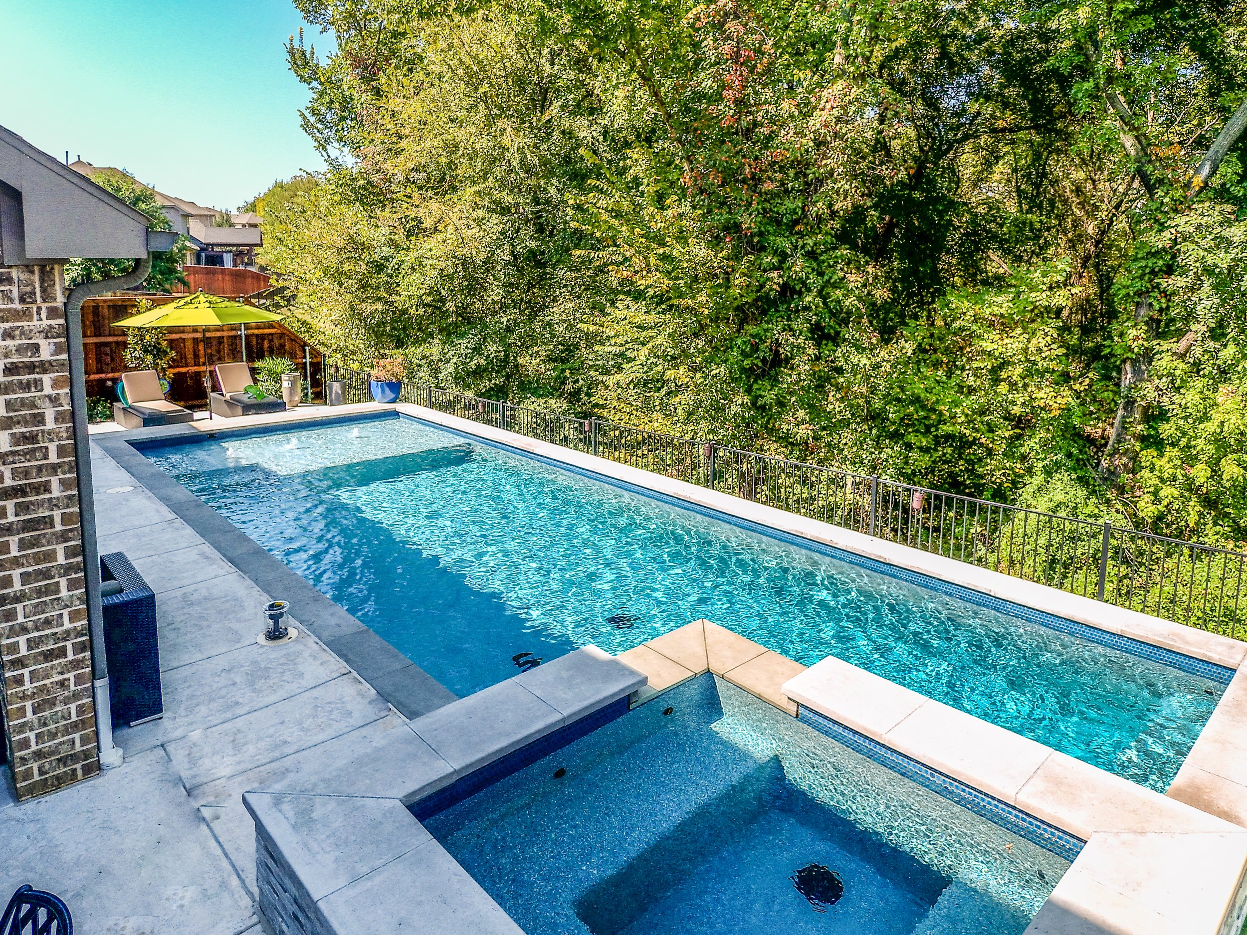 A backyard with a swimming pool and hot tub, surrounded by trees, with outdoor seating and an umbrella.