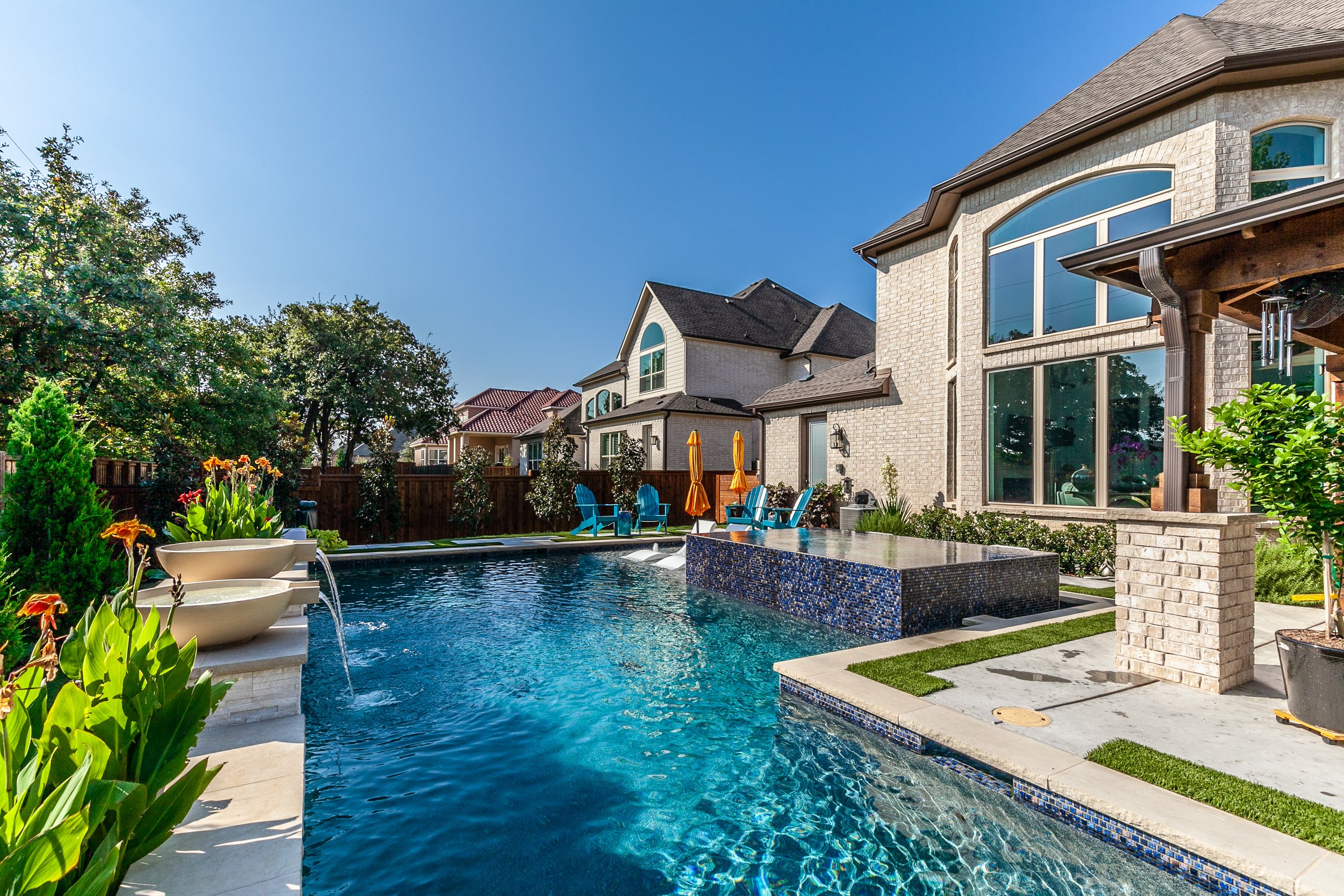 Backyard with a swimming pool, blue chairs, umbrellas, and a waterfall feature, surrounded by a wooden fence and neighboring houses, under a clear blue sky.