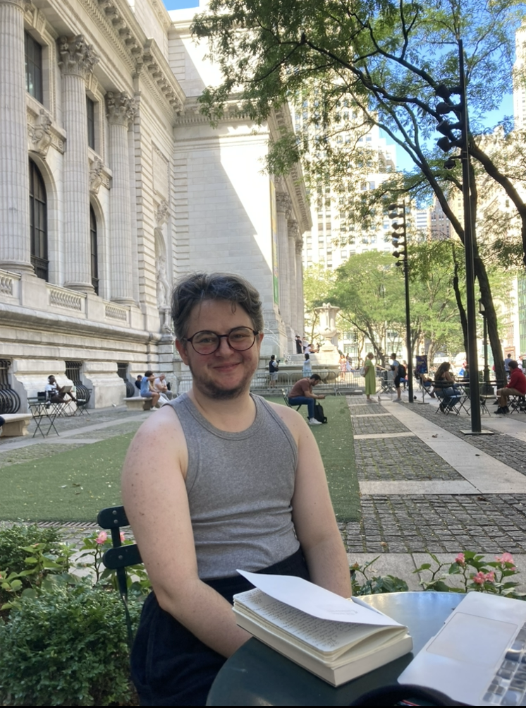 A person with glasses and short hair, wearing a sleeveless gray shirt, sitting at an outdoor table with a book and papers, in front of a historic building with columns, in a park or plaza surrounded by trees and other people.