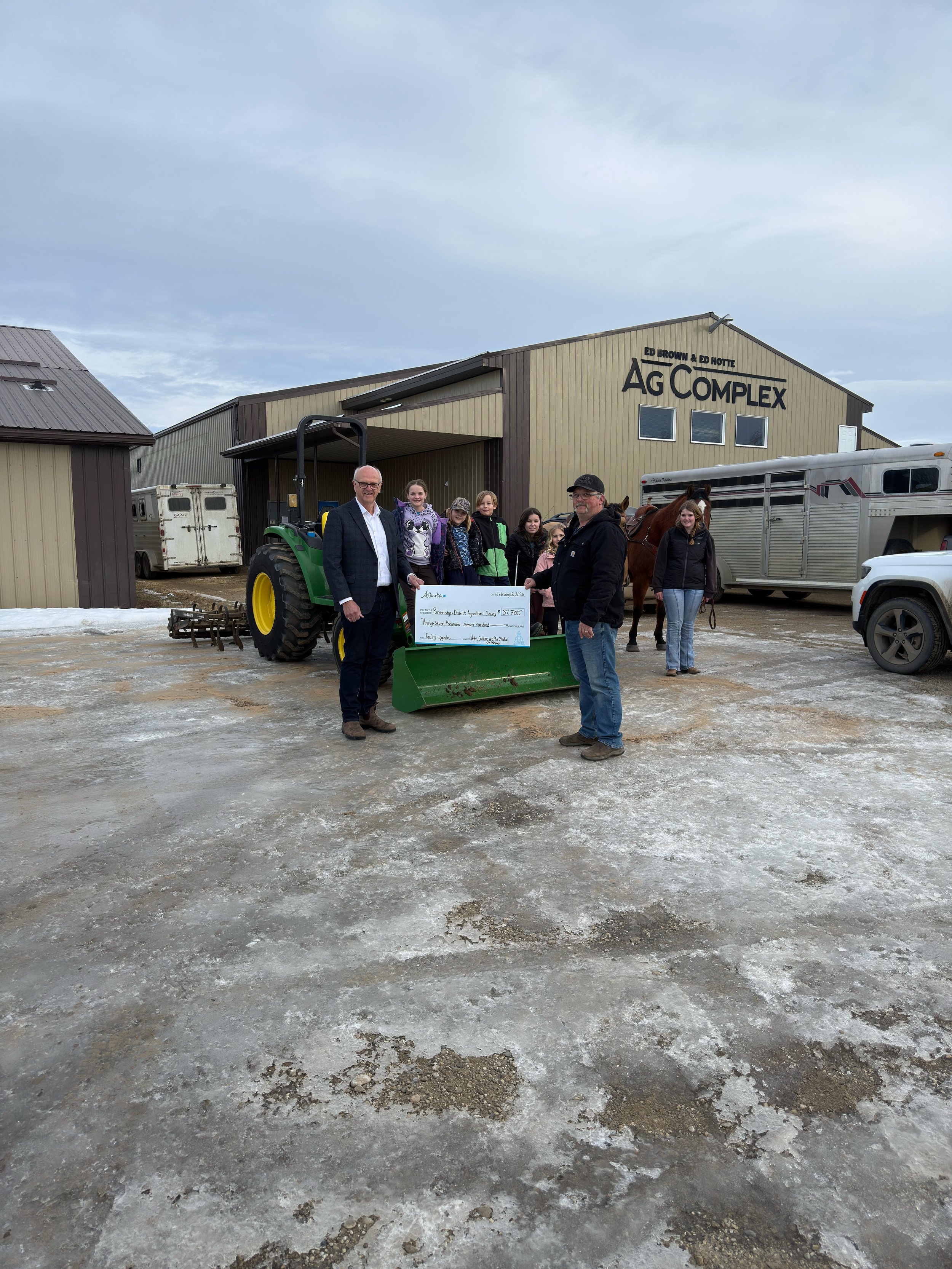 A group of people, including children and adults, standing in front of a barn with a tractor, holding a large check. The barn has signs that say 'Ag Complex' and names 'Ed Brown and Ed Hotte.' One man is holding a check and the others are gathered around, some with horses. The ground is icy and muddy, and the sky is overcast.