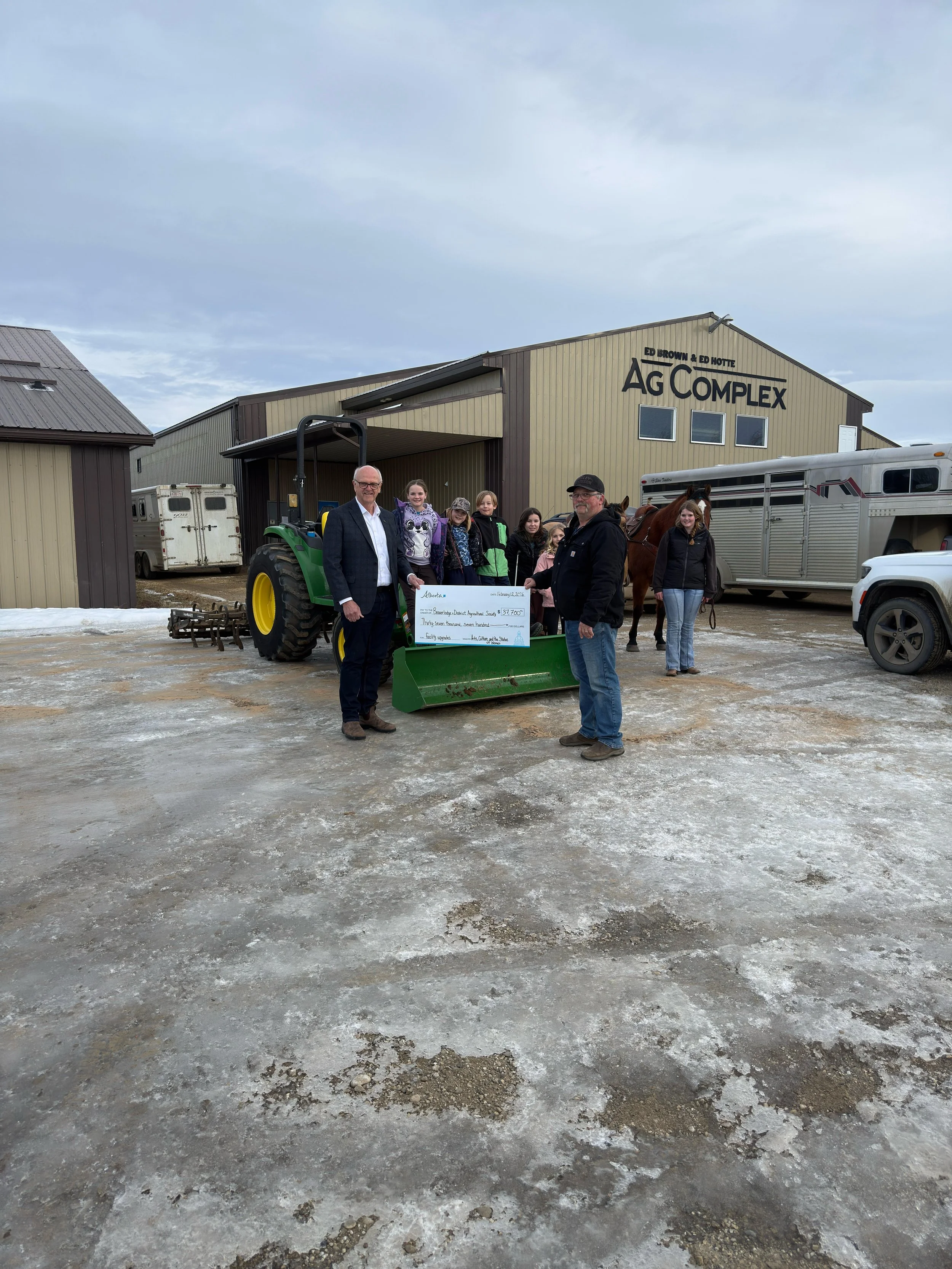 Group of people standing outside a barn labeled 'AG COMPLEX' with a large check and a tractor, likely during a donation or award event.