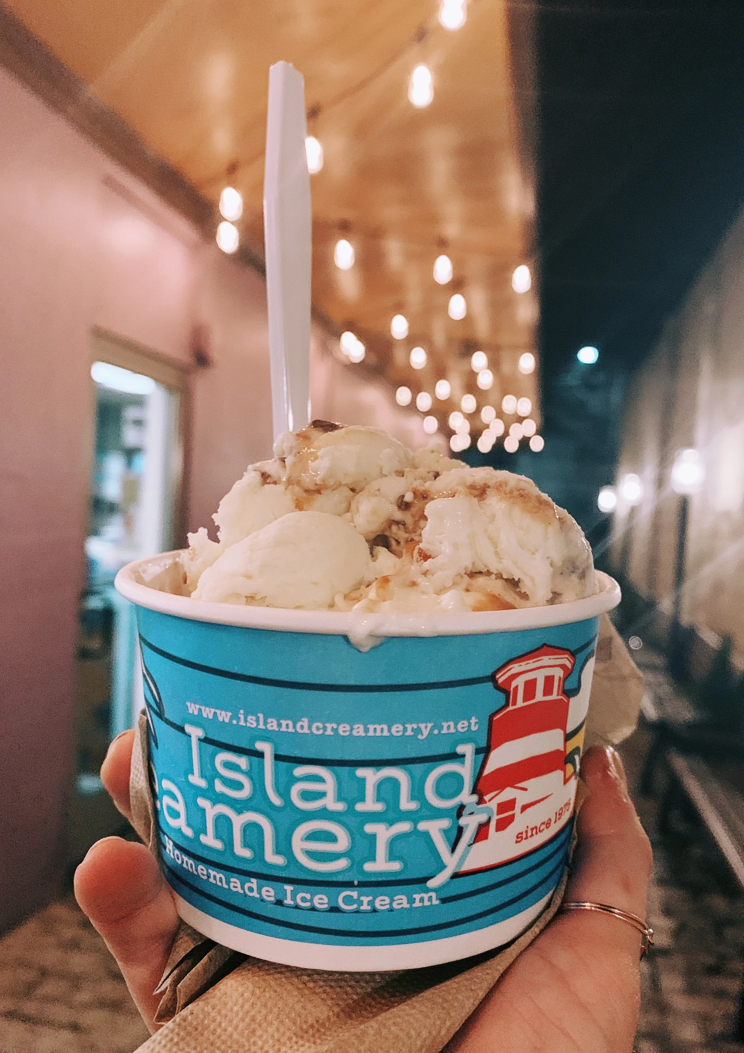 A cup of Island Creamery ice cream with a spoon in it, held by a person in a warm brown paper napkin, with a background of hanging lights indoors.