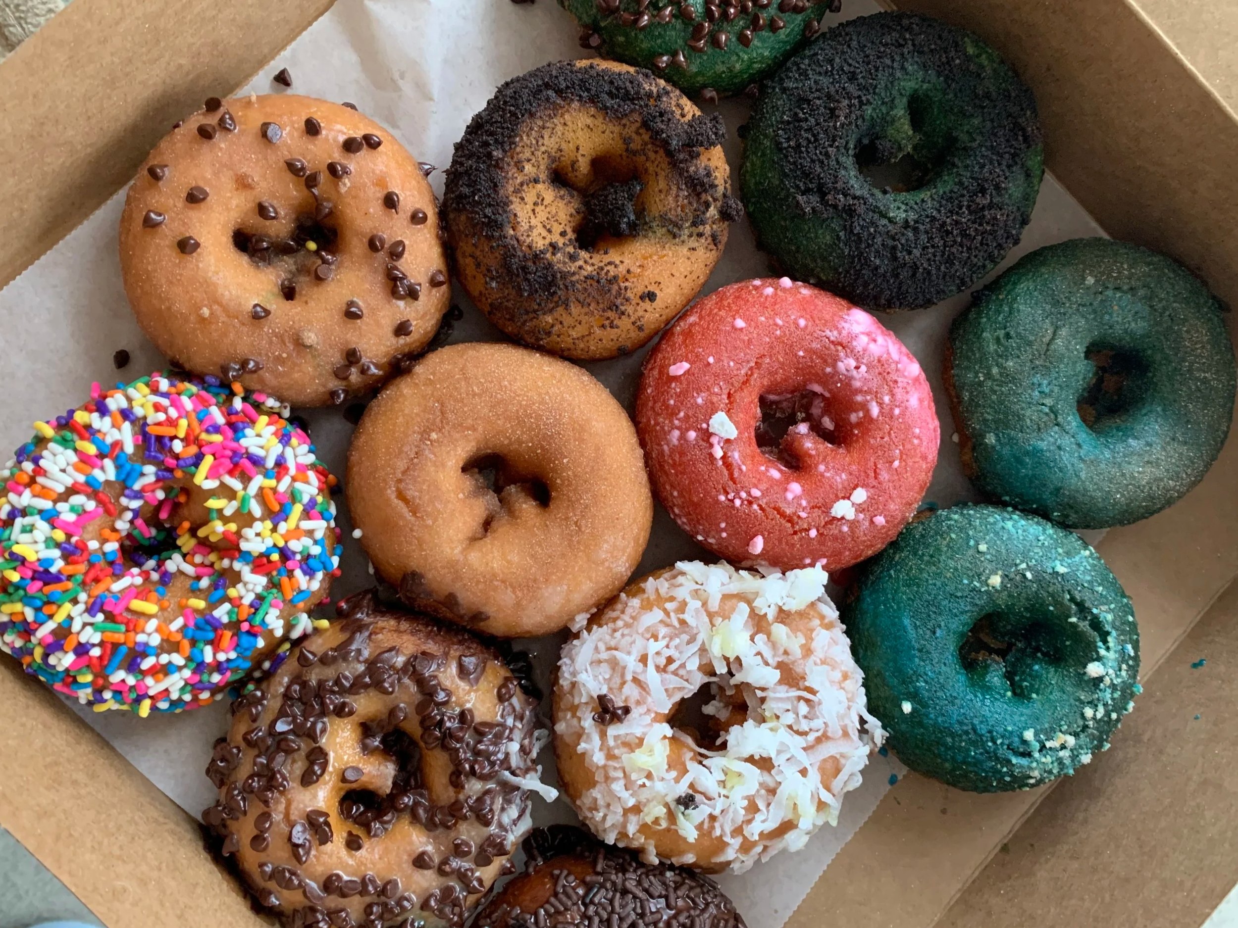 Assorted donuts with different toppings, including rainbow sprinkles, chocolate chips, coconut flakes, and colored sugar, arranged in a box.