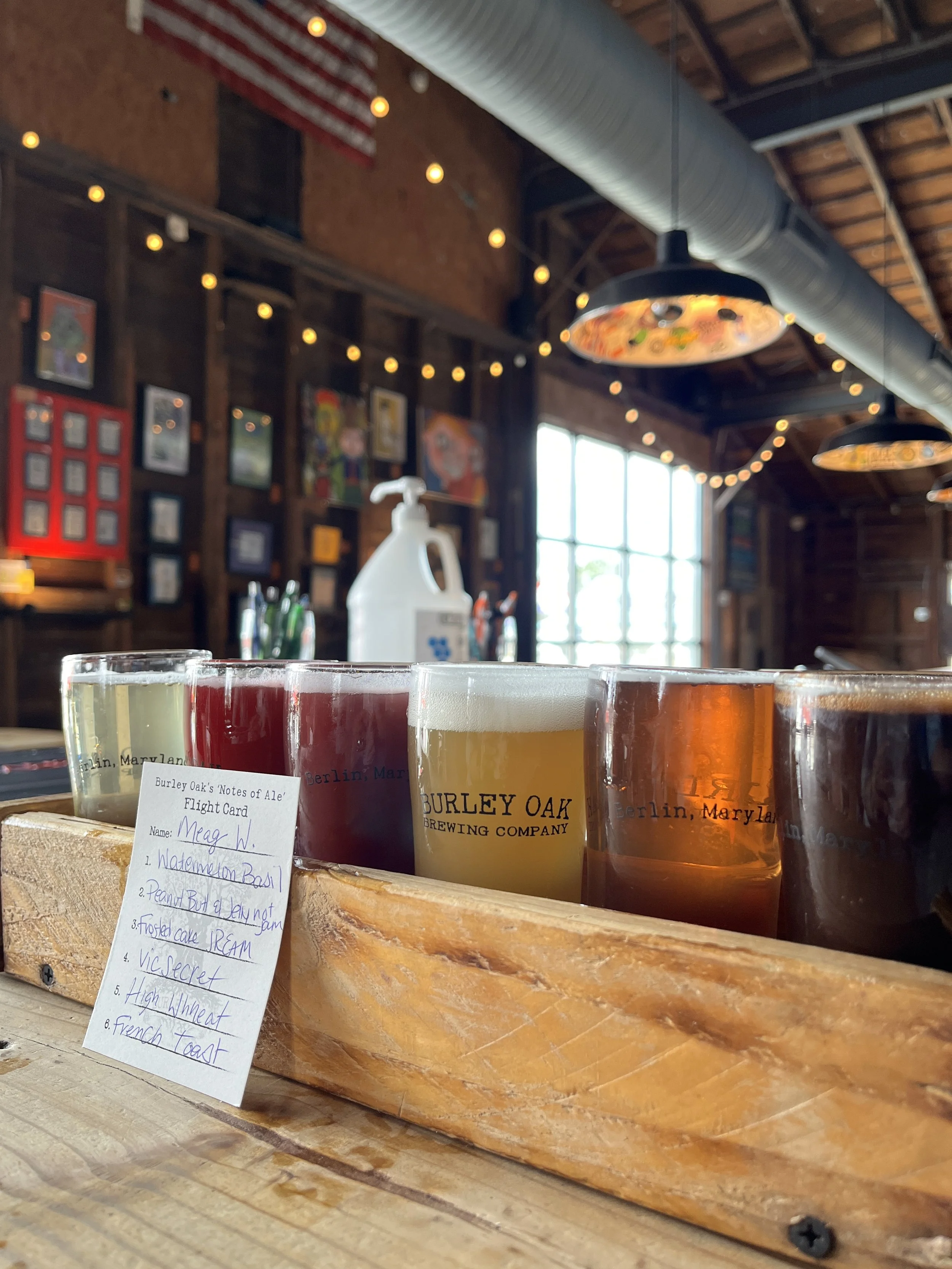 A wooden tray with four tasting glasses of beer from Burley Oak Brewing Company, each with different colors, placed on a rustic wood surface. A handwritten menu is in front of the tray listing beer flight choices.