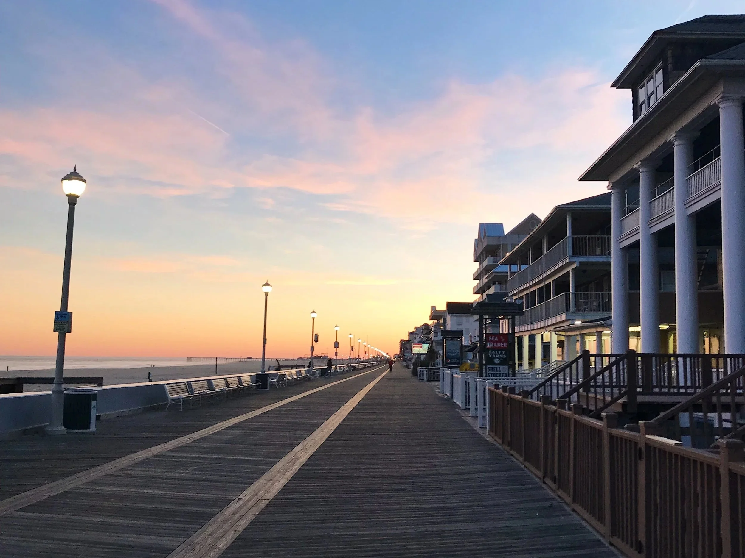 Sunset view on the boardwalk of Ocean City Maryland with lamps and a row of beach houses on the right side, overlooking the ocean and sky with pink and blue clouds.