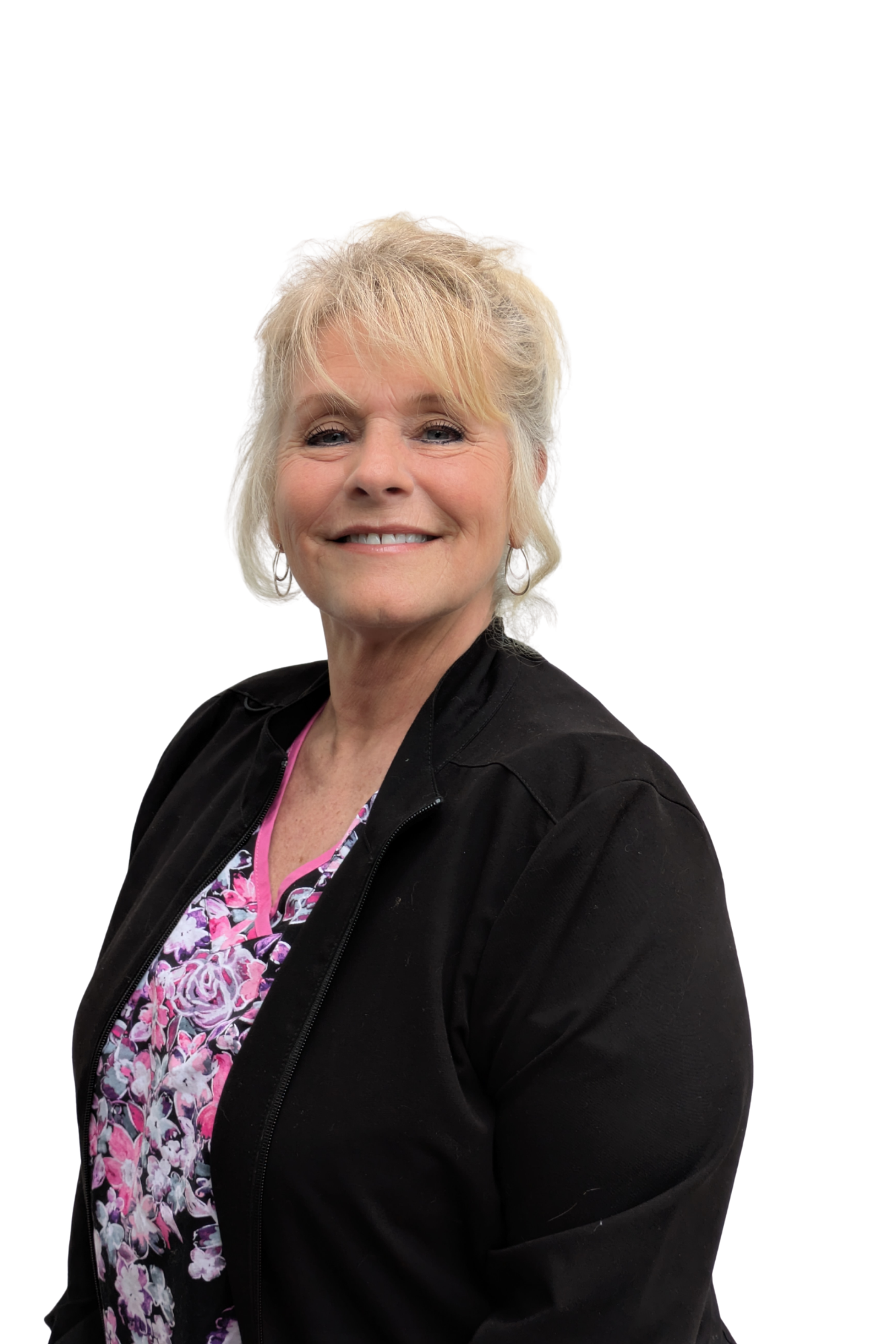 A woman with short, blonde, slightly messy hair, smiling and wearing a black jacket over a pink and purple floral top, standing against a plain white background.