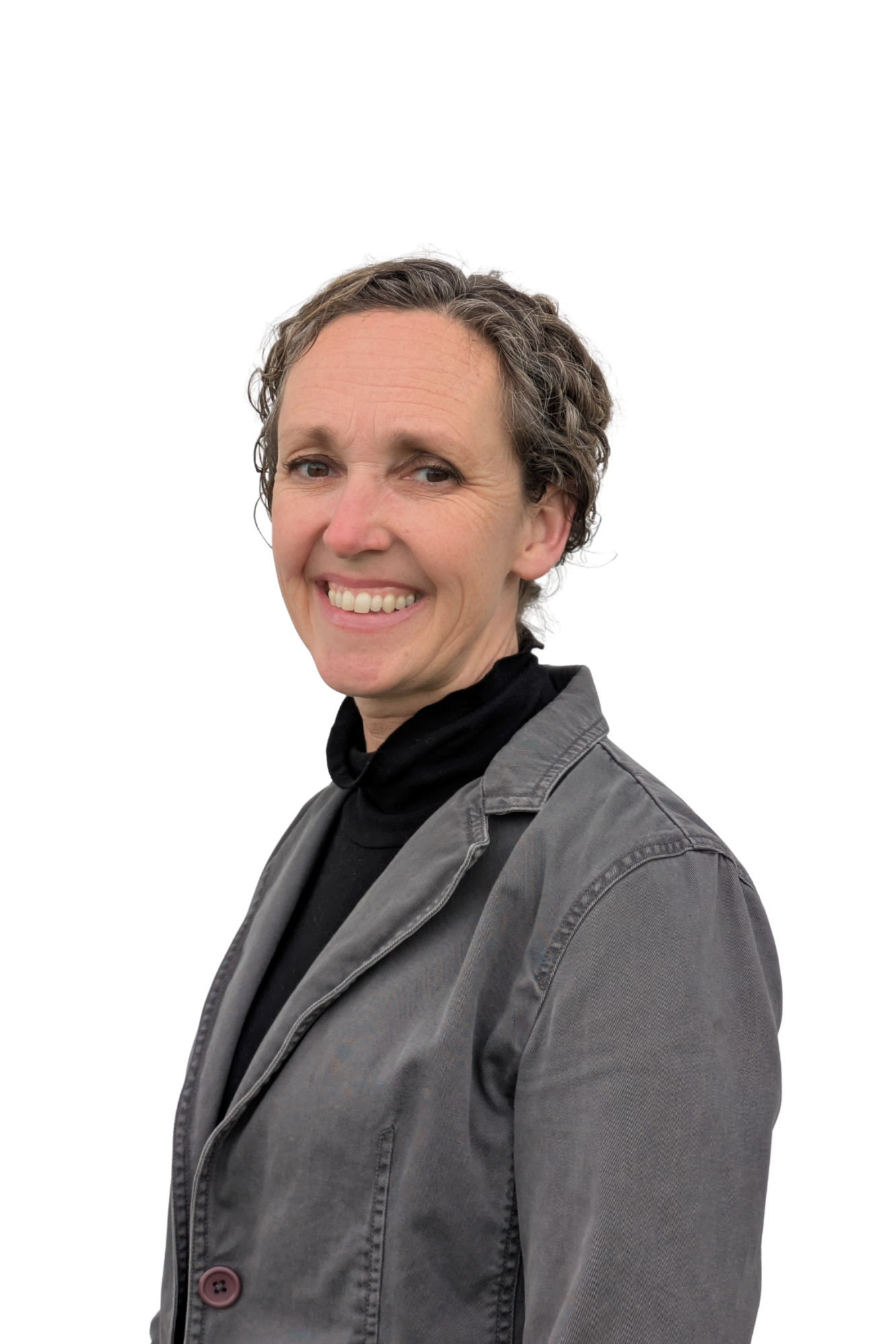 A woman with short curly gray hair smiling, wearing a black turtleneck, gray jacket, and standing against a white background.