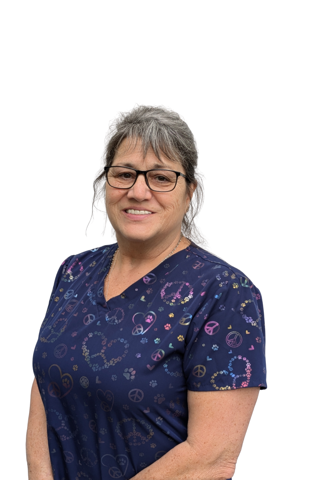 A middle-aged woman with gray hair, wearing glasses and a navy blue scrub top decorated with peace signs and colorful hearts, smiling against a plain white background.