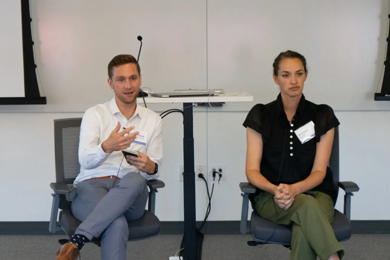 A man and a woman sitting in chairs during a discussion or presentation in a conference room. The man is speaking, holding a smartphone, wearing a white shirt and gray pants, and the woman is listening attentively, wearing a black blouse and green pants.