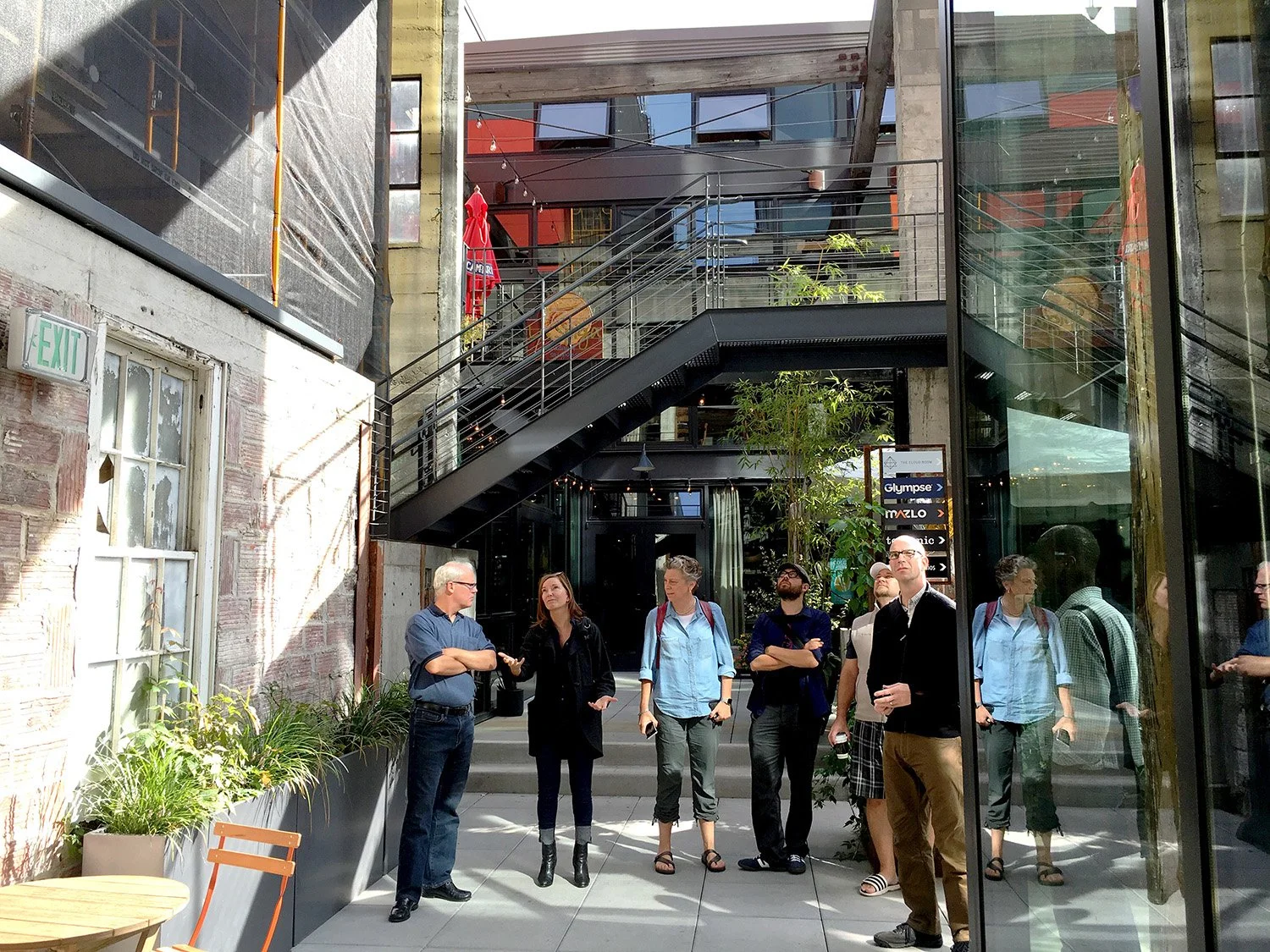 Group of people standing and talking outside a modern building with glass walls, stairs, and plants, during daytime.