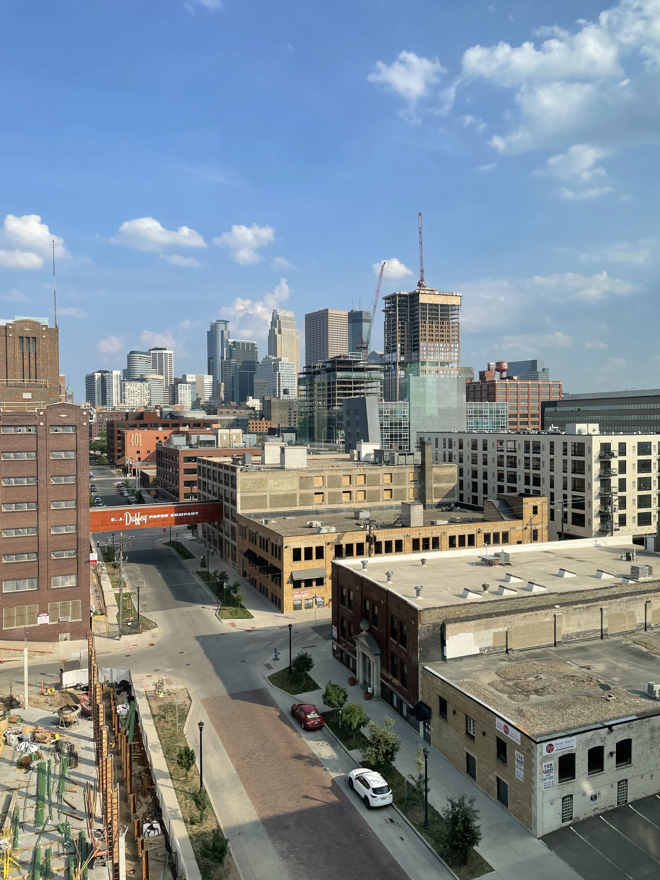 A cityscape with skyscrapers under a blue sky with scattered clouds, including buildings of varying heights and architectural styles. Some buildings are under construction with cranes visible.